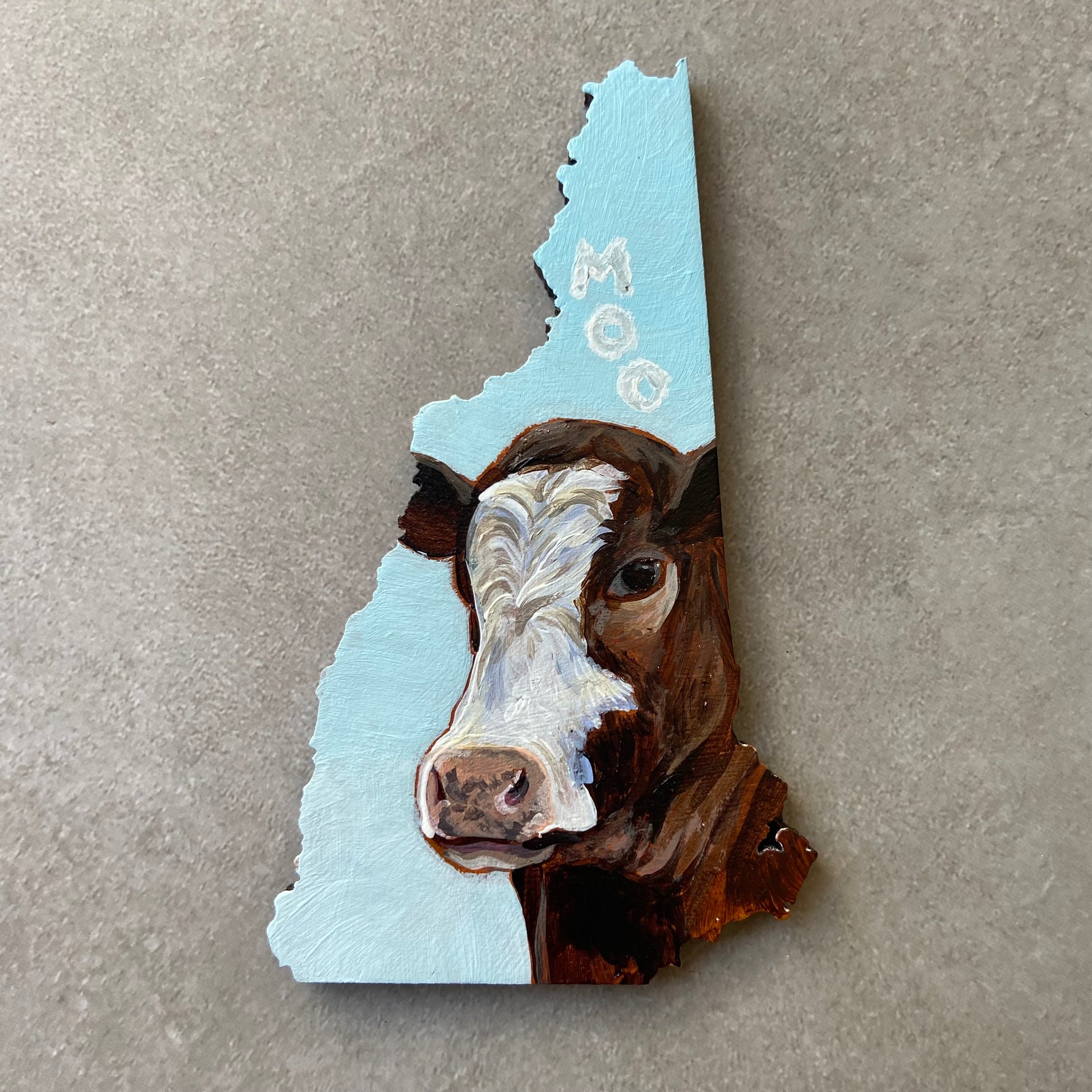 A New Hampshire-shaped painting depicting a brown and white cow's face looking at the viewer. The sky-blue background has three letter-shaped clouds over the cow's head, reading "MOO".