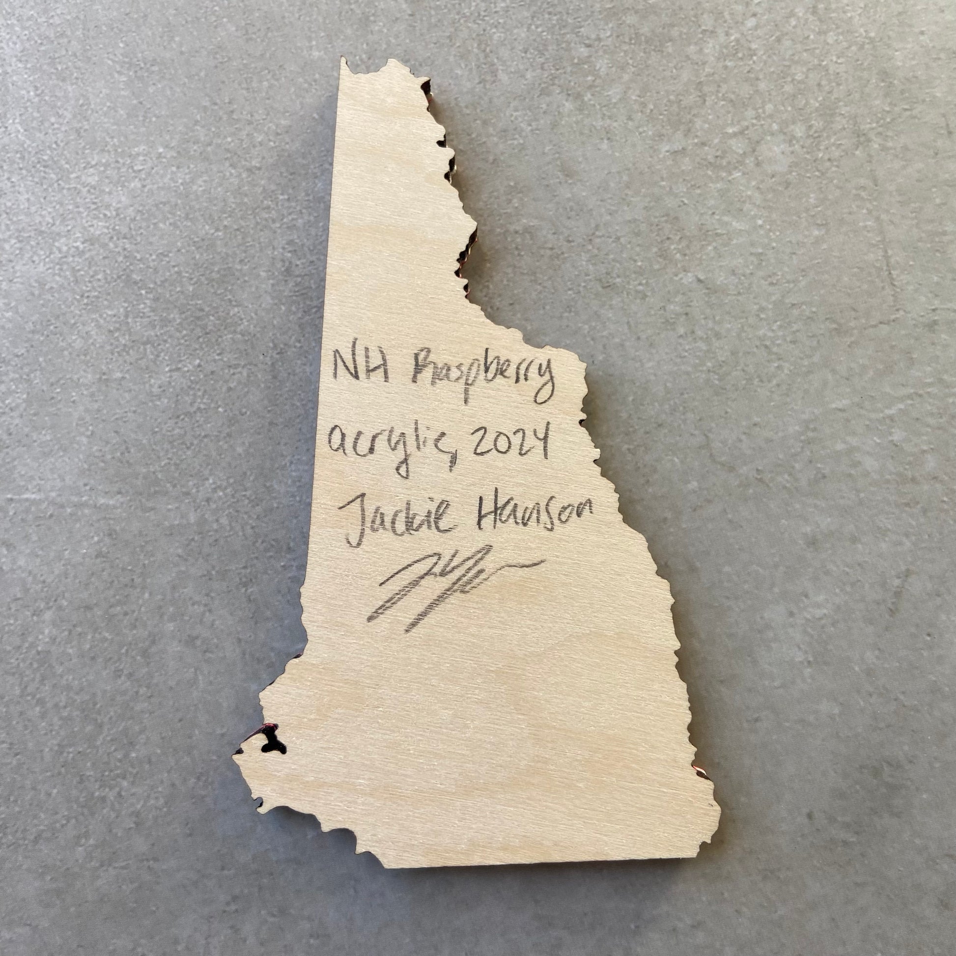 The back of a New Hampshire-shaped painting on wood. Writing on the bare wood in pencil reads, "NH Raspberry. Acrylic, 2024. Jackie Hanson" and signed by the artist.