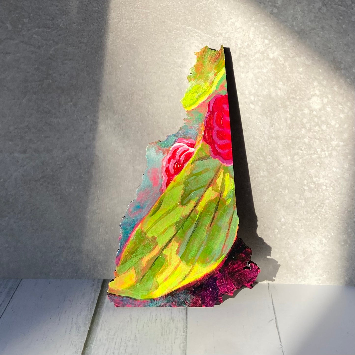 A New Hampshire-shaped painting showing a sunlit green leaf with part of two ripe raspberries visible. The painting is propped upright in a patch of sun.