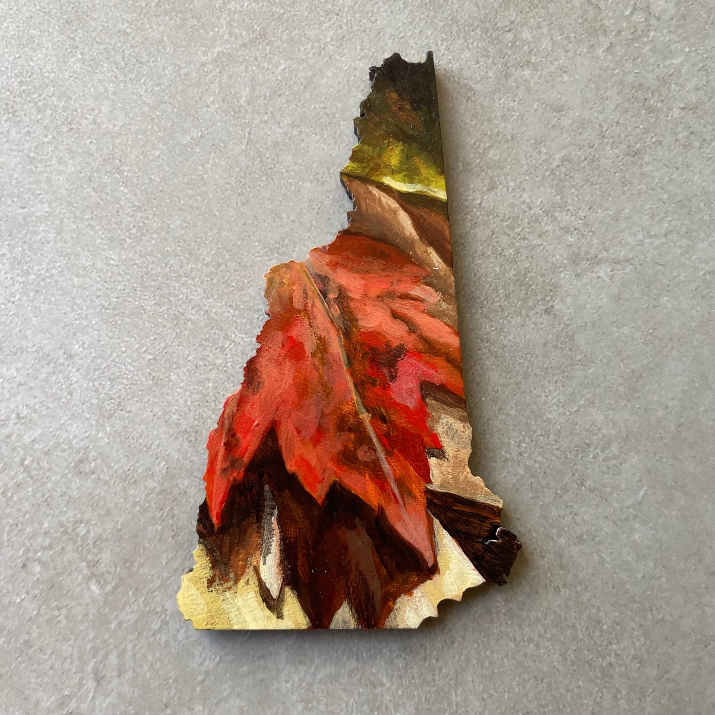 A New Hampshire-shaped painting depicting a red maple leaf on the ground among other fall leaves.