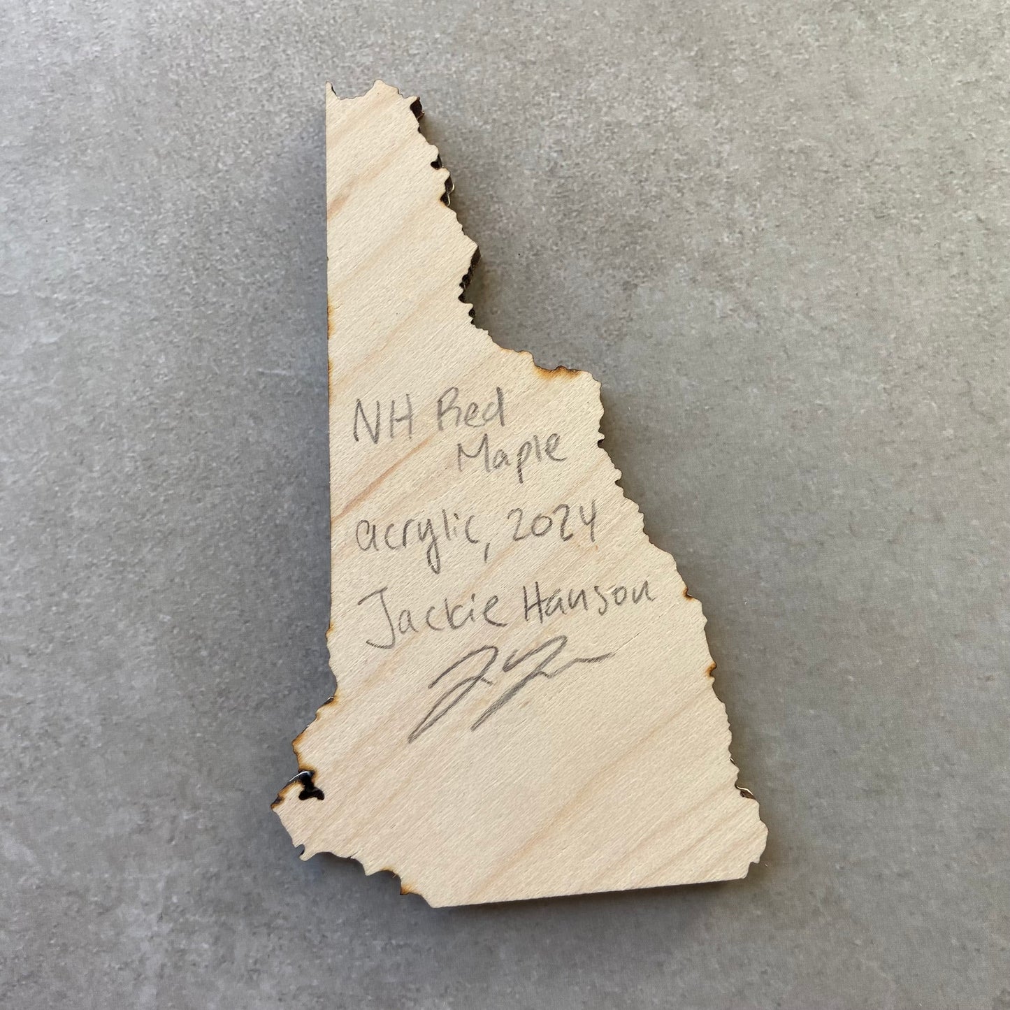 The back of a New Hampshire-shaped painting on wood. Writing on the bare wood in pencil reads, "NH Red Maple. Acrylic, 2024. Jackie Hanson" and signed by the artist.