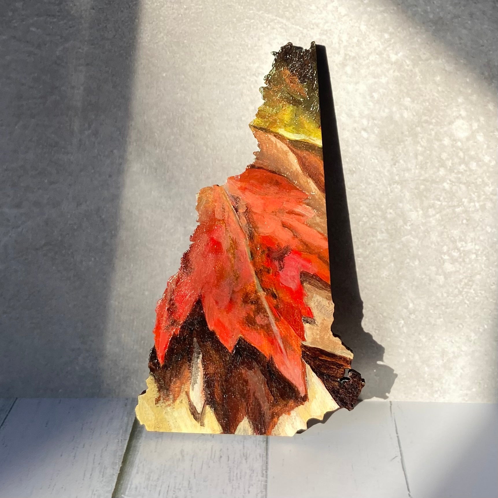A New Hampshire-shaped painting depicting a red maple leaf on the ground among other fall leaves. The painting is propped up in a sunny spot.