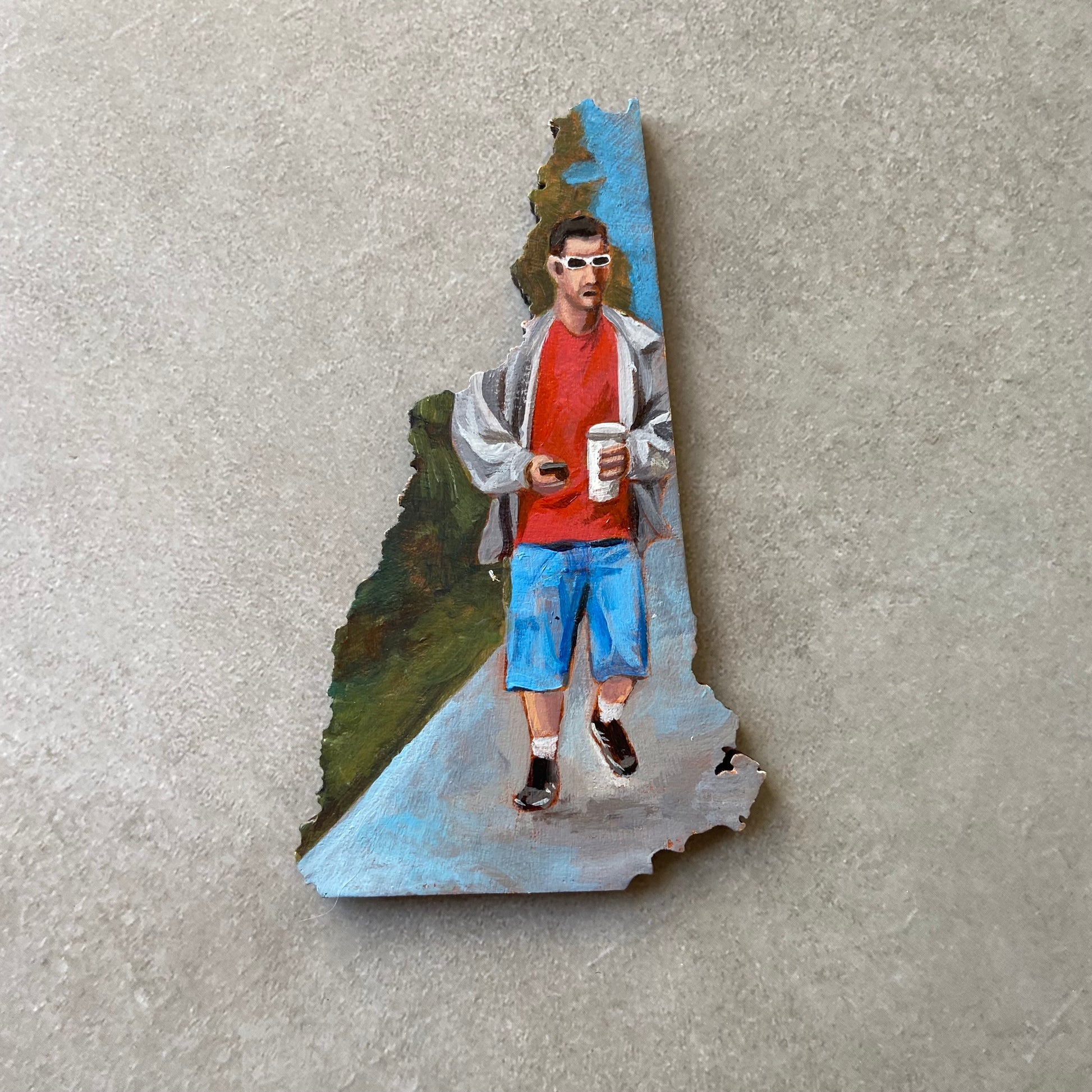 A New-Hampshire shaped painting of Adam Sandler in a classic Sandler outfit. He is walking on a sidewalk carrying a cell phone in one hand and a disposable coffee cup in the other.
