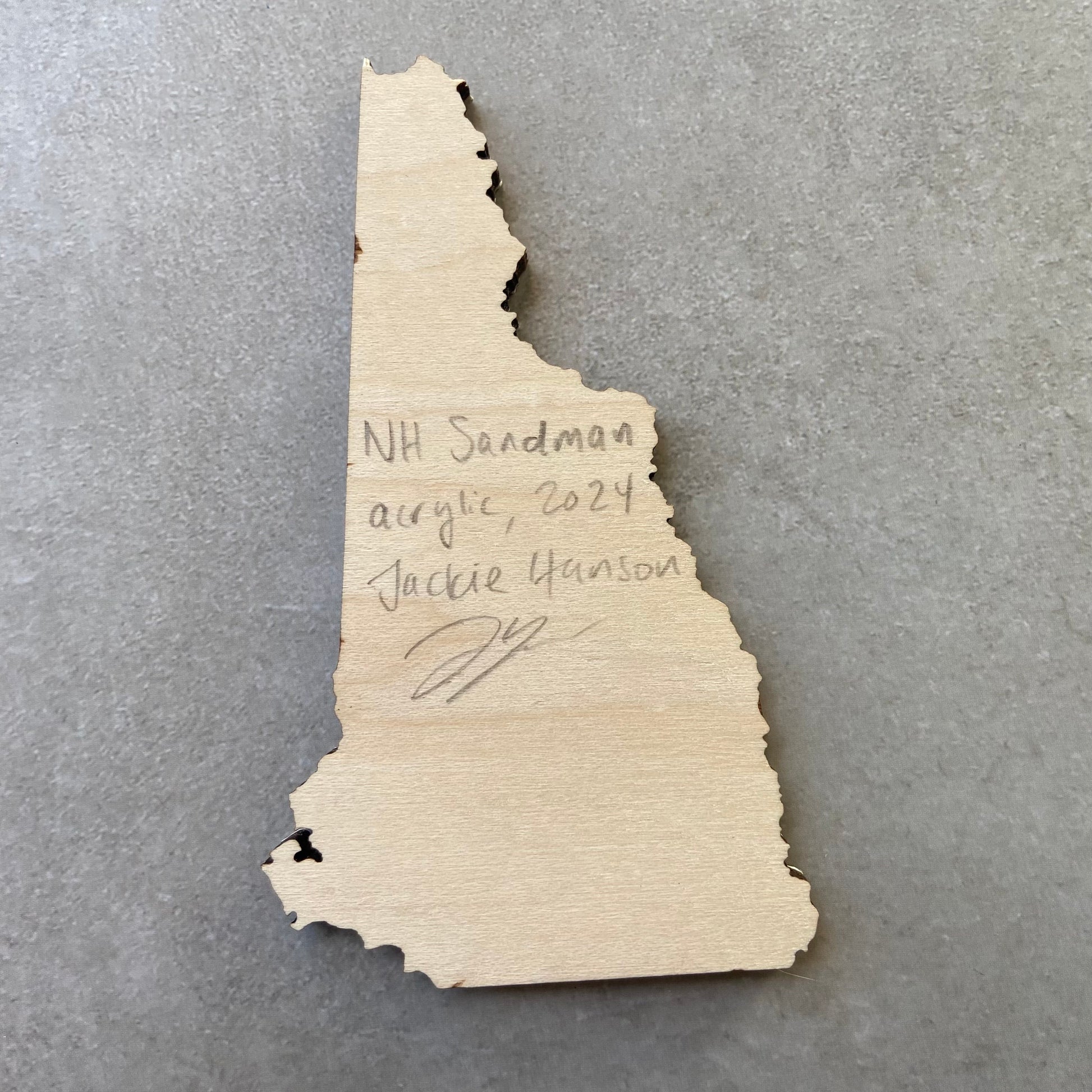 The back of a New Hampshire-shaped painting on wood. Writing on the bare wood in pencil reads, "NH Sandman. Acrylic, 2024. Jackie Hanson" and signed by the artist.