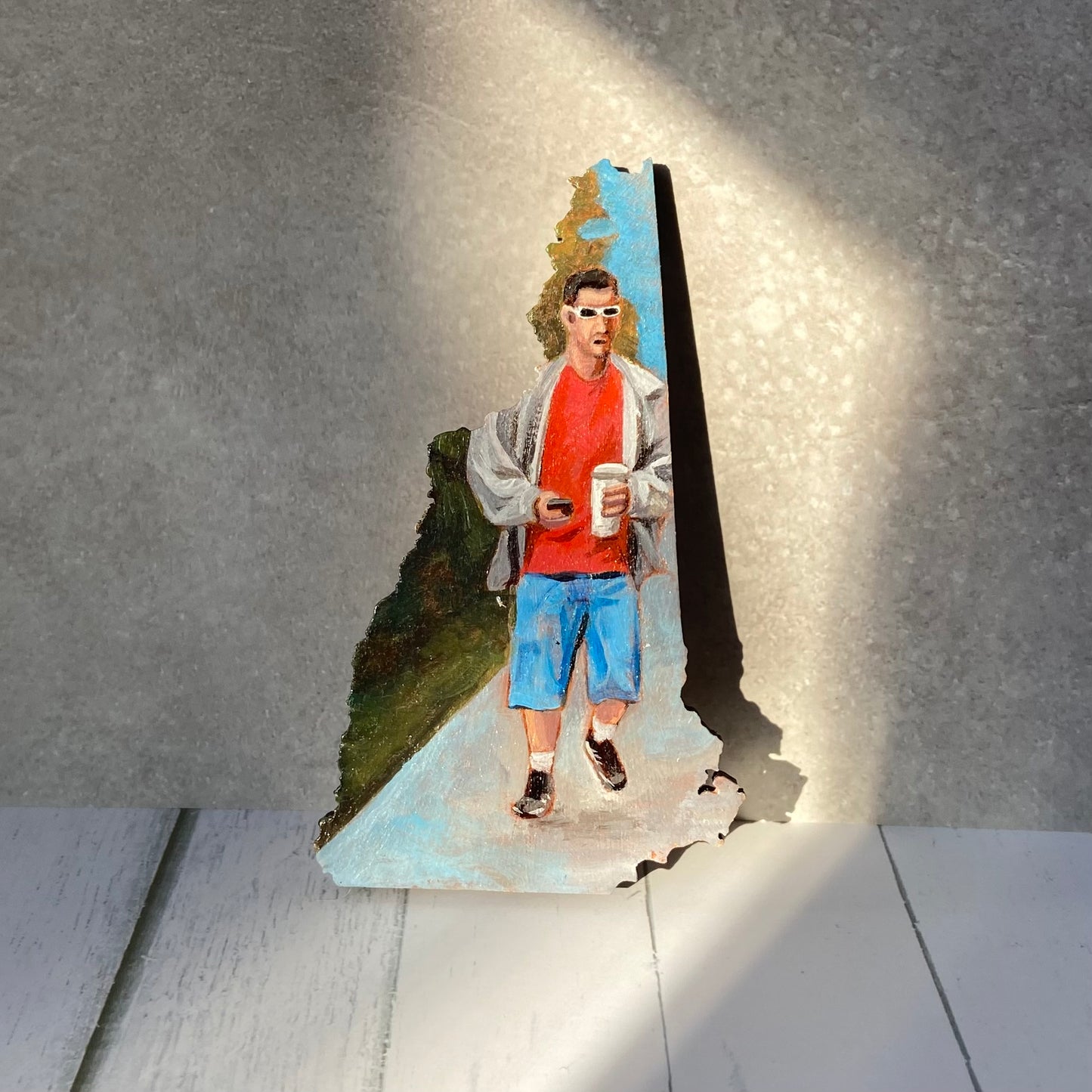 A New-Hampshire shaped painting of Adam Sandler in a classic Sandler outfit. He is walking on a sidewalk carrying a cell phone in one hand and a disposable coffee cup in the other. The painting is upright in a patch of sunlight.