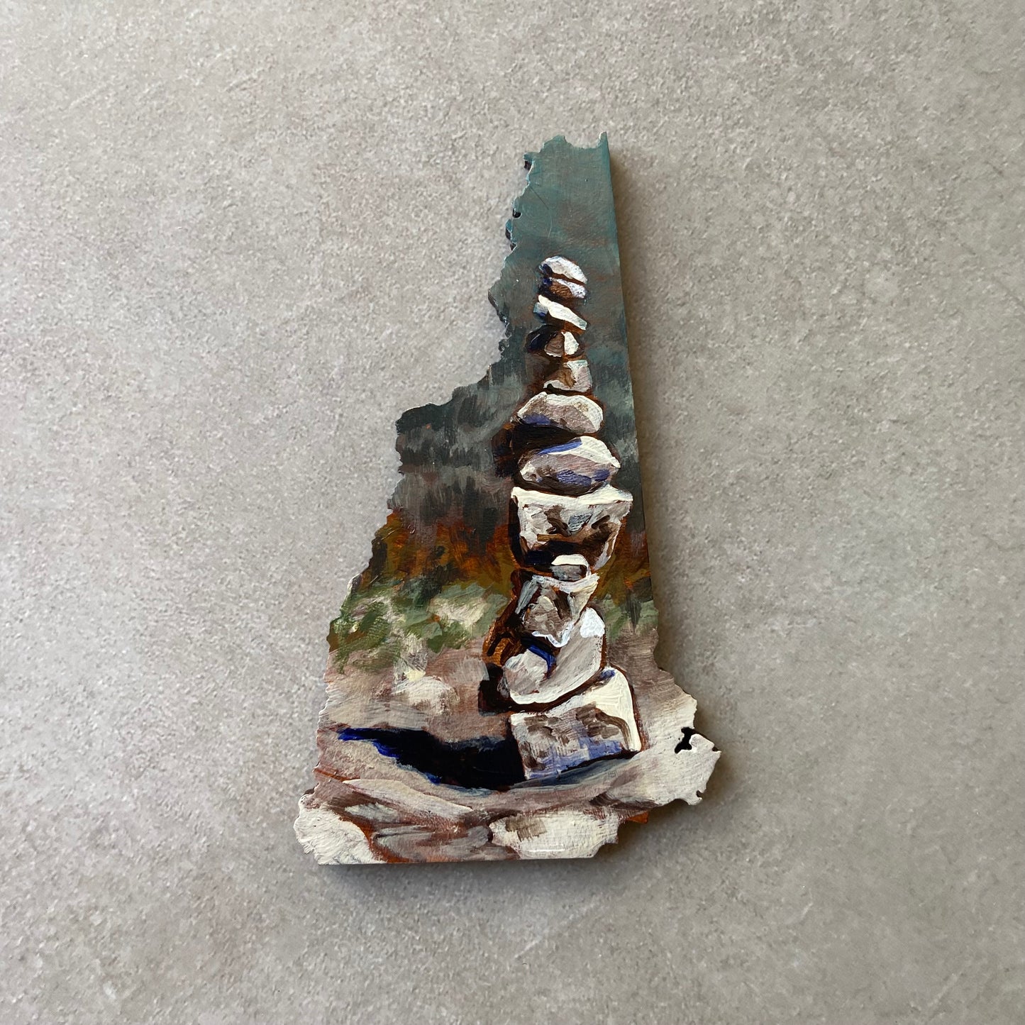 A New Hampshire-shaped painting of stacked rocks on a rock surface, with implied forest painted in the distance.
