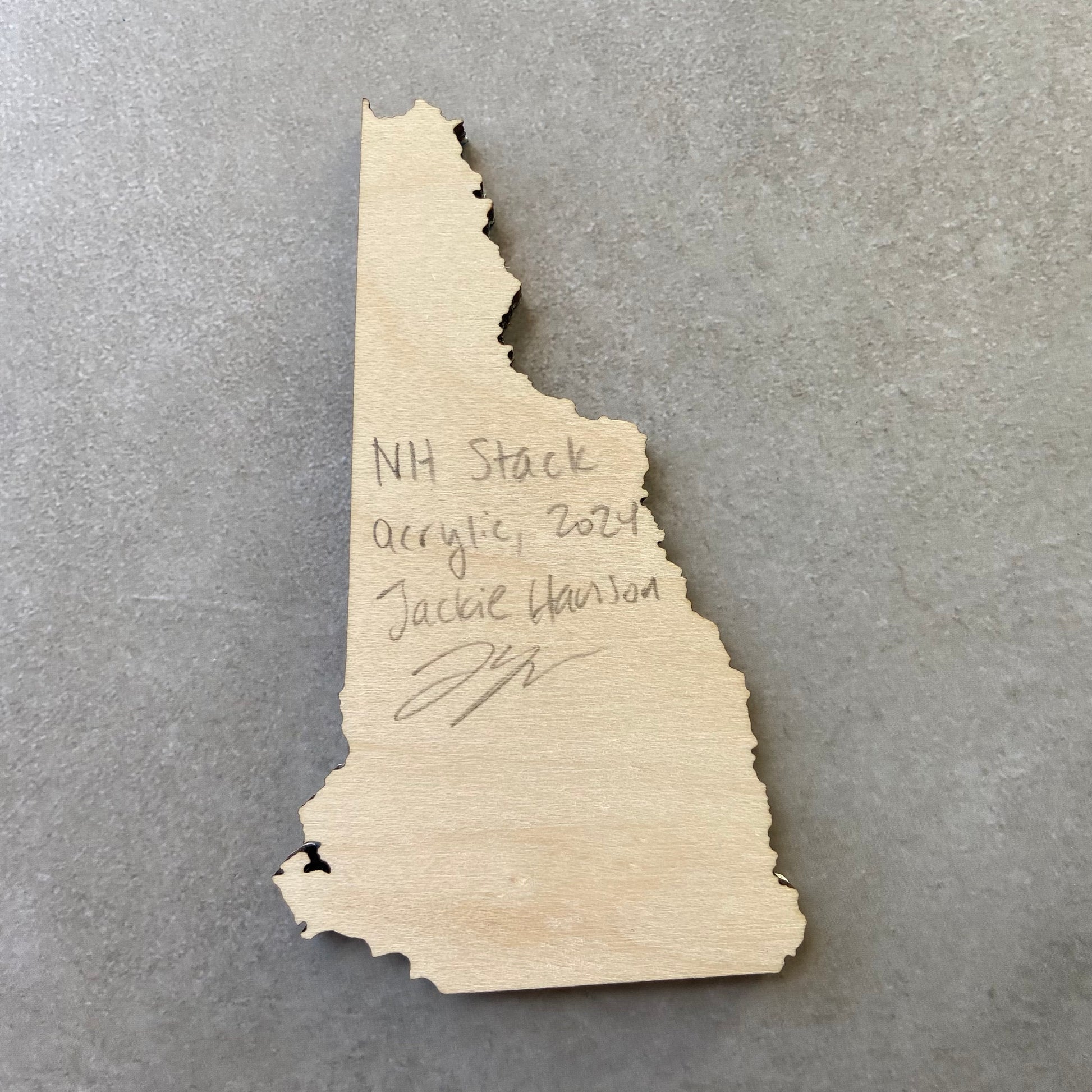 The back of a New Hampshire-shaped painting on wood. Writing on the bare wood in pencil reads, "NH Stack. Acrylic, 2024. Jackie Hanson" and signed by the artist.