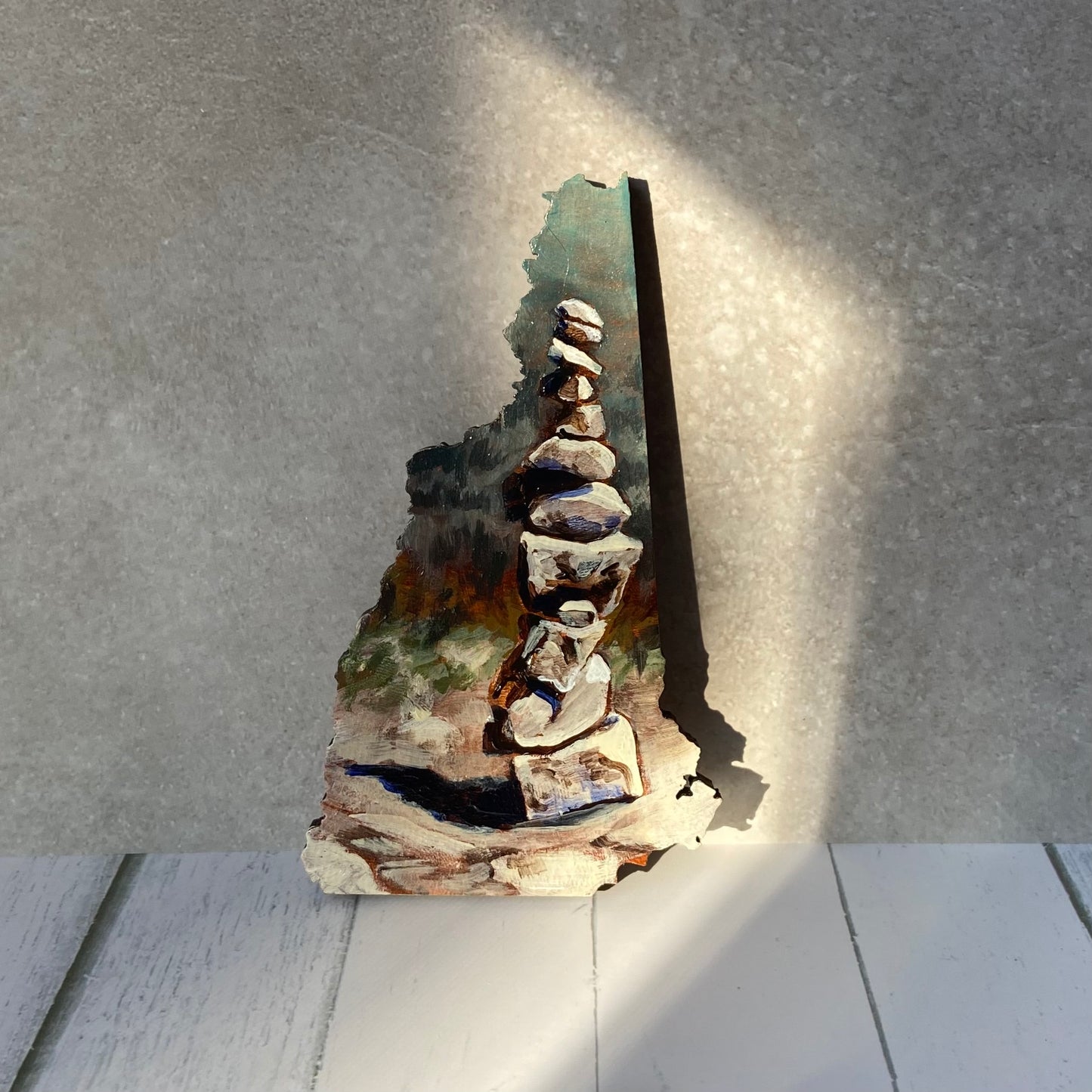 A New Hampshire-shaped painting of stacked rocks on a rock surface, with implied forest painted in the distance. The painting is upright in a patch of sun.