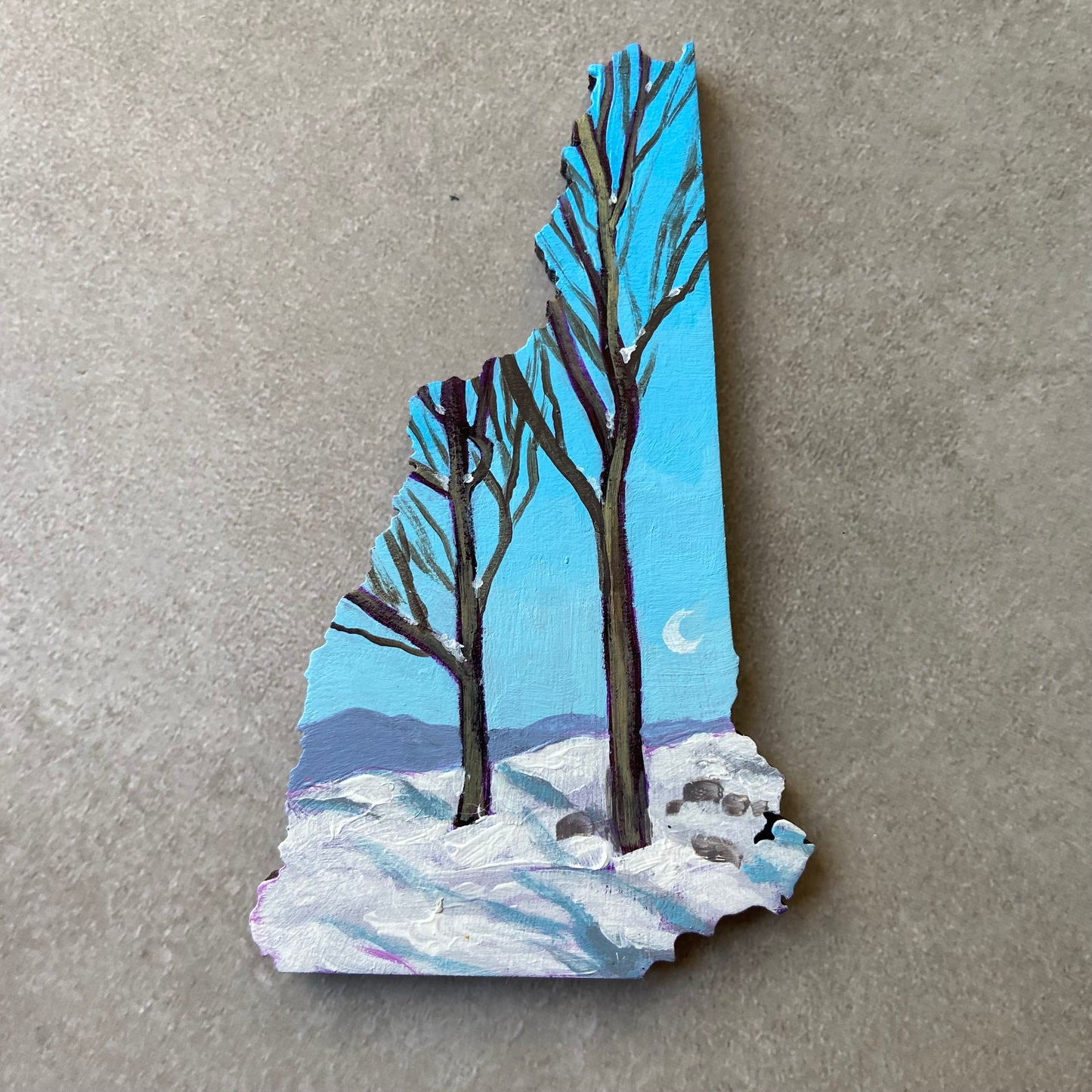 A New Hampshire-shaped painting of two bare-branched trees in the snow with mountains in the distance. The sky is clear, blue, and has a crescent moon at right.