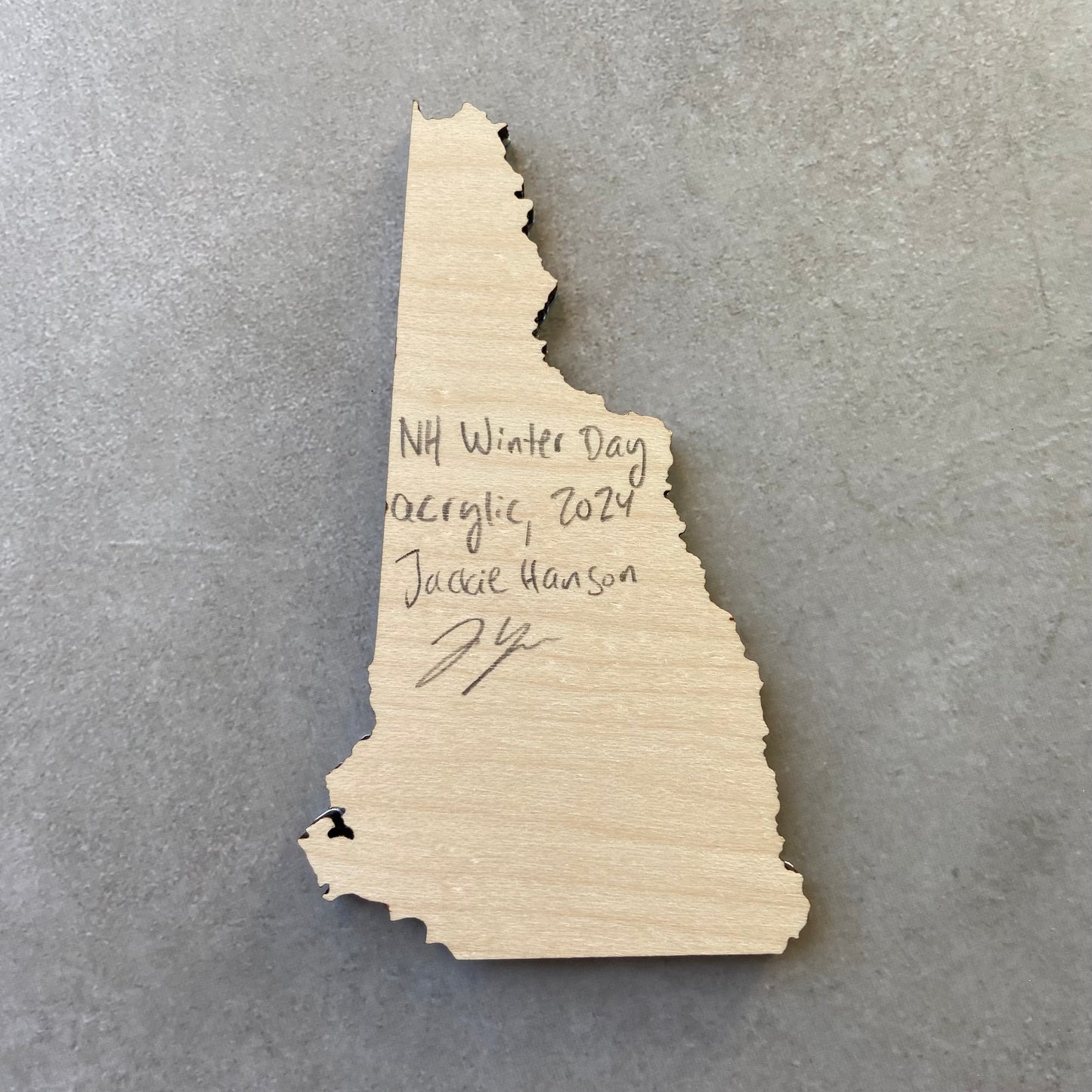 The back of a New Hampshire-shaped painting on wood. Writing on the bare wood in pencil reads, "NH Winter Day. Acrylic, 2024. Jackie Hanson" and signed by the artist.