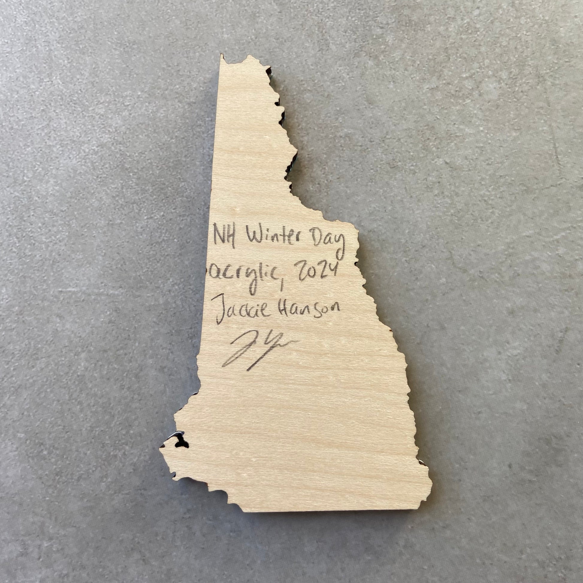 The back of a New Hampshire-shaped painting on wood. Writing on the bare wood in pencil reads, "NH Winter Day. Acrylic, 2024. Jackie Hanson" and signed by the artist.