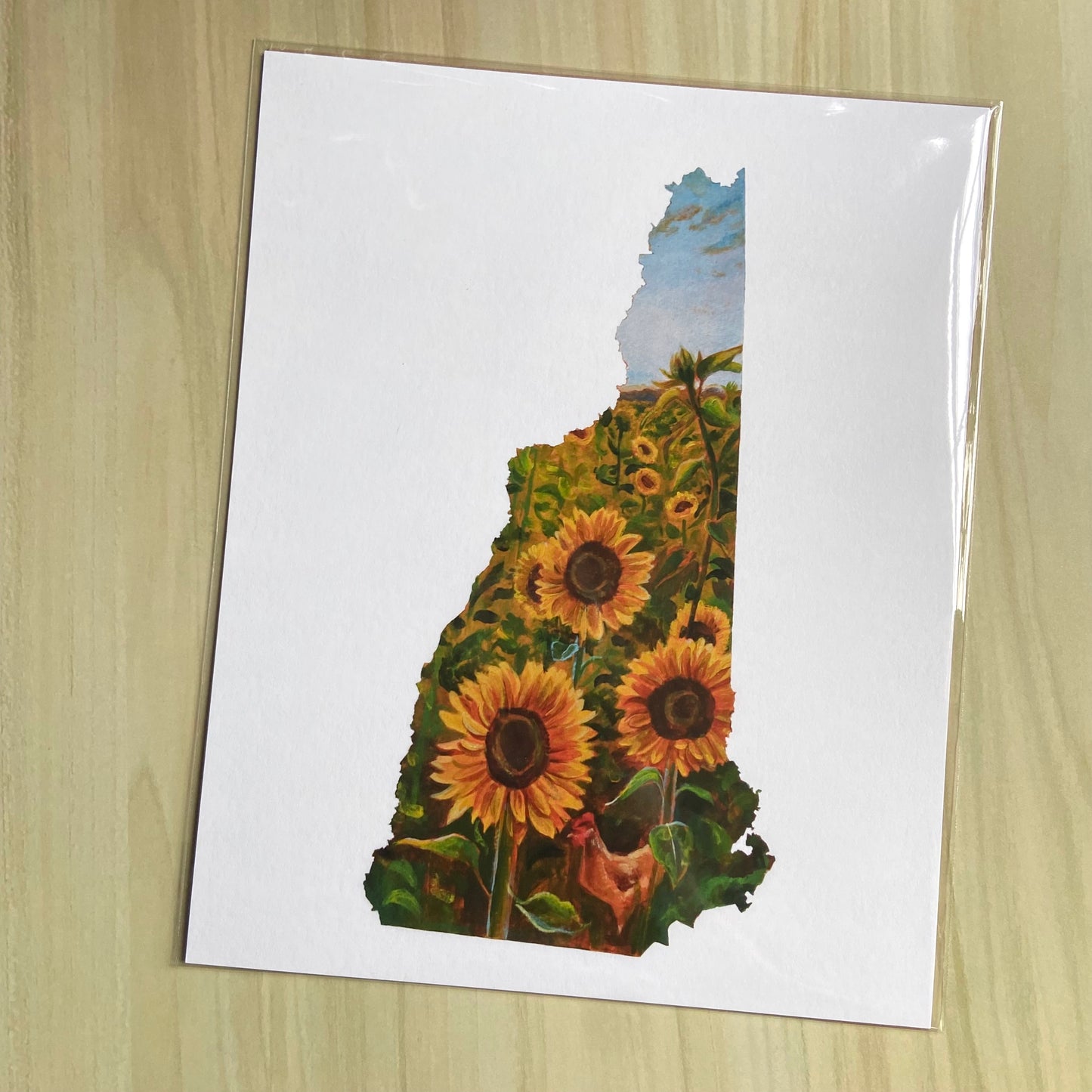 An art print on paper of a sunflower field painting in the shape of New Hampshire. Outside the map outline shape is white. The focus is three yellow sunflowers and an orange hen near the bottom. More flowers are farther behind and the sky is blue.