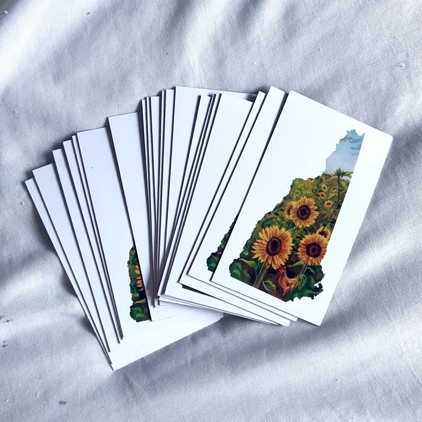 A pile of rectangular white magnets with a painting of sunflowers in the shape of New Hampshire.