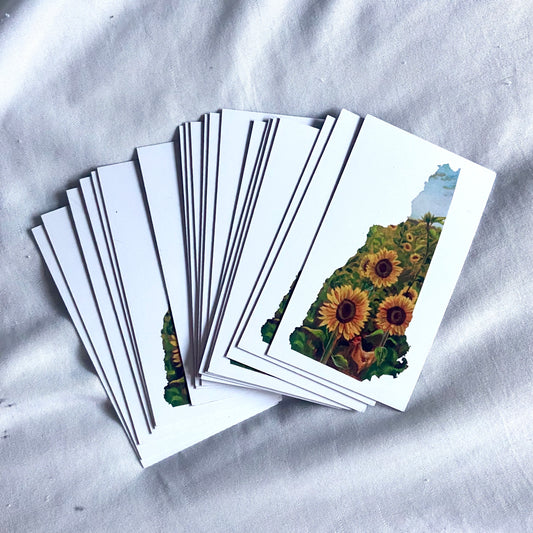 A pile of rectangular white magnets with a painting of sunflowers in the shape of New Hampshire.