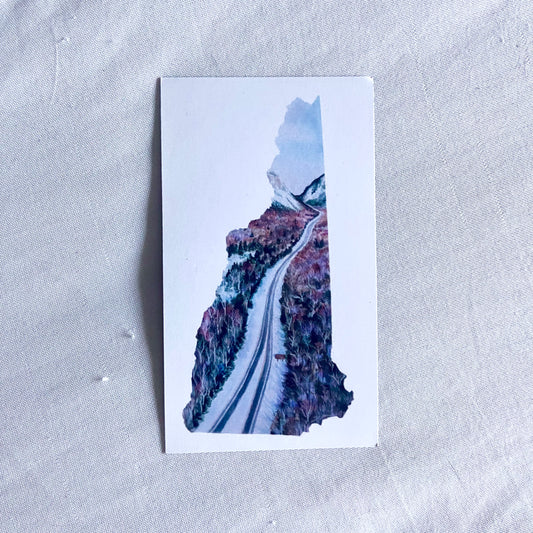 A rectangular magnet with a blue painting in the shape of New Hampshire and white around it. The painting shows a snowy divided highway moving up through the woods to a valley between two mountains.