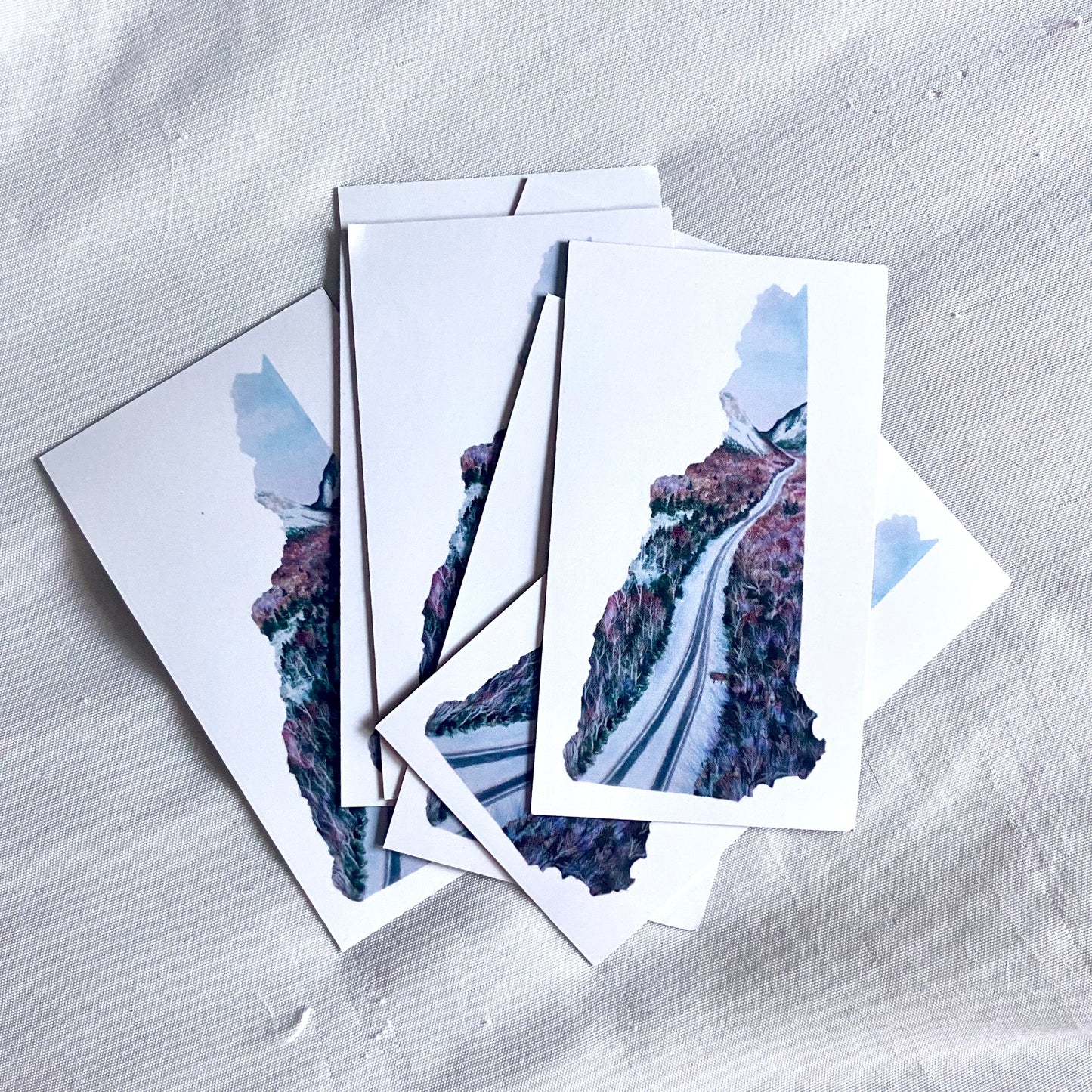 A pile of rectangular magnets, each white with a painting in the shape of New Hampshire in the center. The artwork is a winter scene of Interstate 93 going through Franconia Notch.