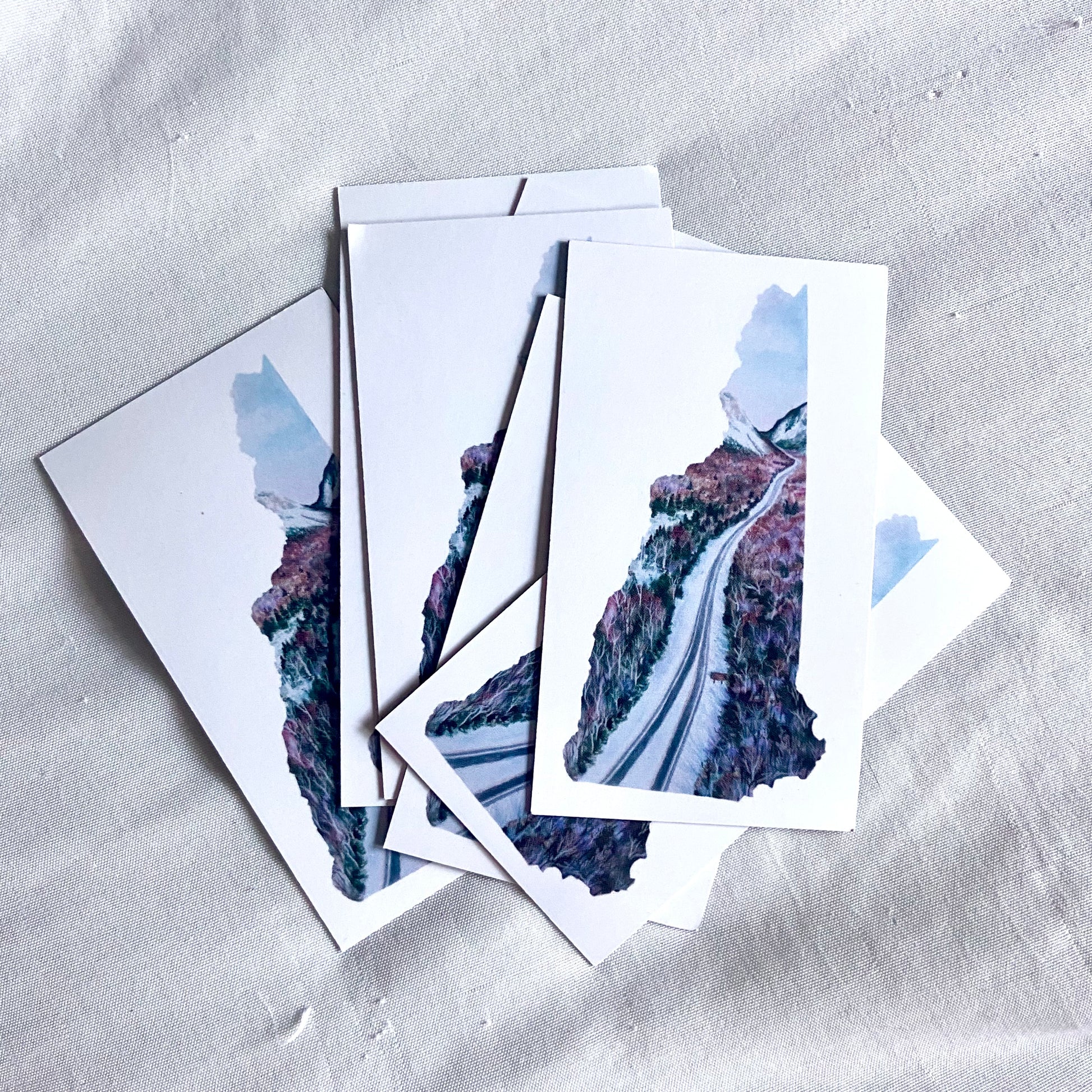 A pile of rectangular magnets, each white with a painting in the shape of New Hampshire in the center. The artwork is a winter scene of Interstate 93 going through Franconia Notch.