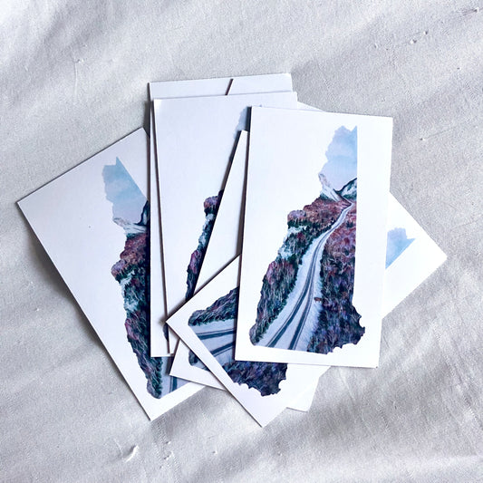 A pile of rectangular magnets, each white with a painting in the shape of New Hampshire in the center. The artwork is a winter scene of Interstate 93 going through Franconia Notch.