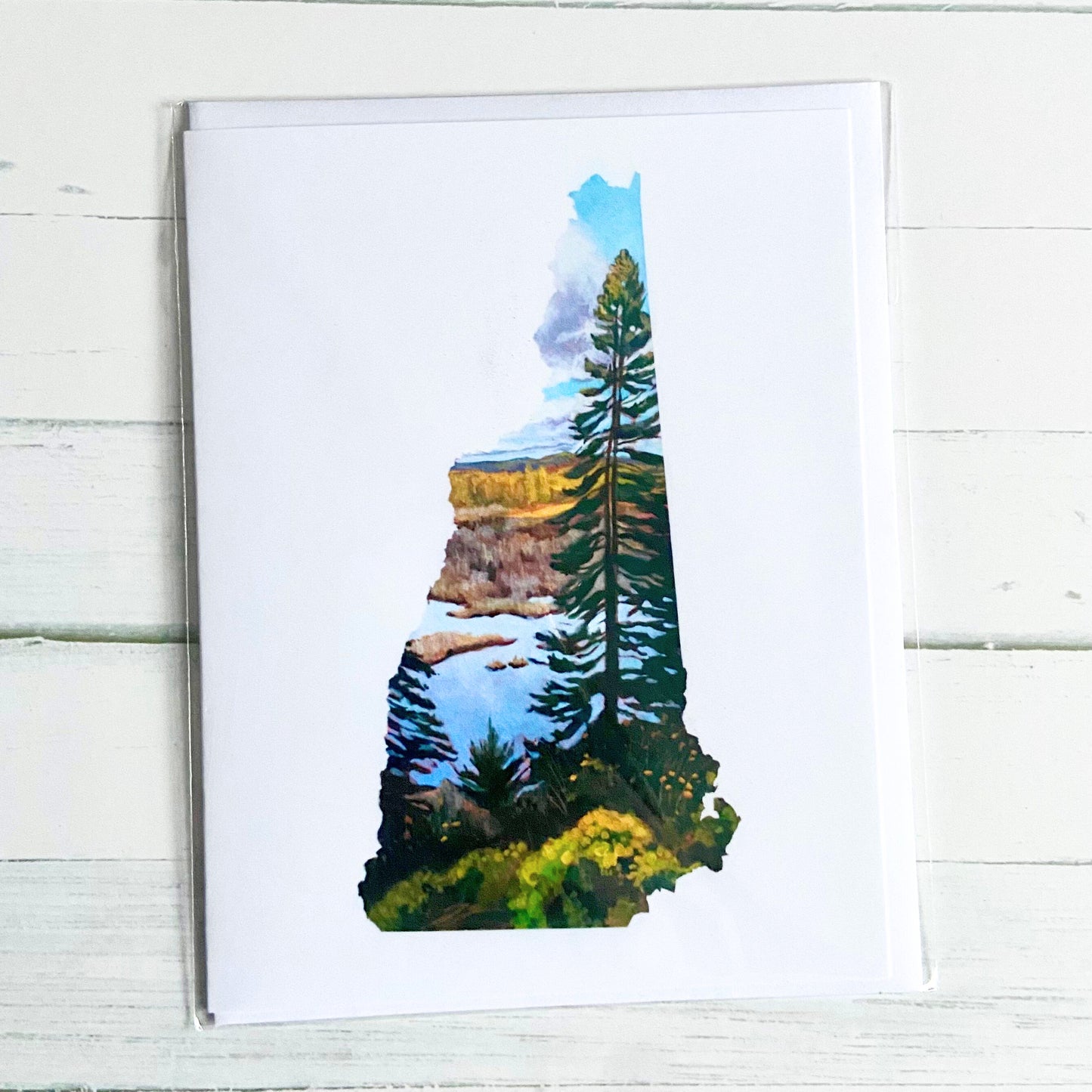 printed on the front. Depicted is a painting in the shape of the state of New Hampshire with a tall pine tree in front of a marsh and fall foliage. The image is on a white background.