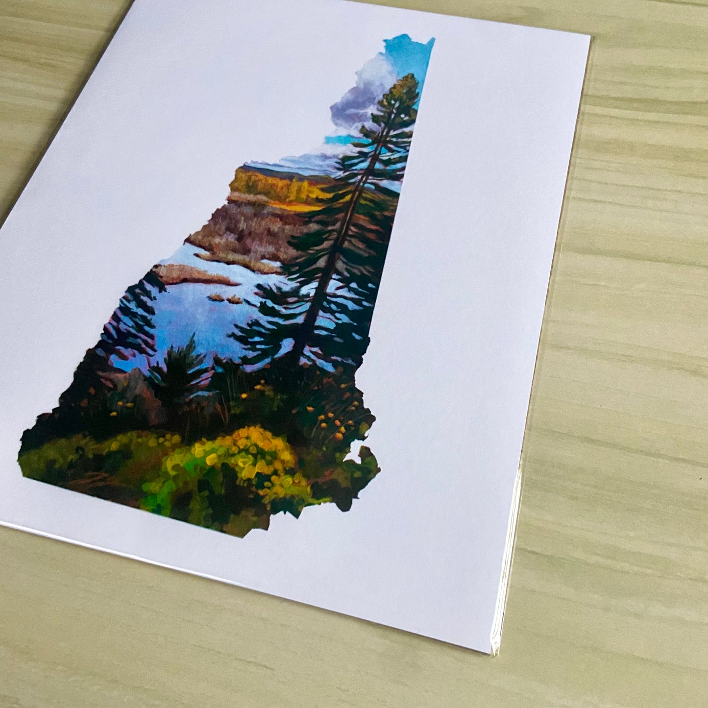 An angled view of a print of my painting, New Hampshire IV. Depicted is a painting in the shape of the state of New Hampshire with a tall pine tree in front of a marsh and fall foliage. The image is on a white background.