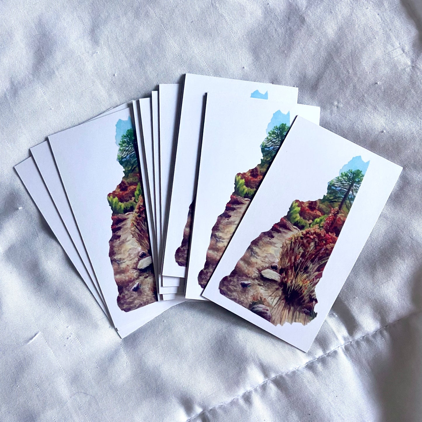 A pile of rectangular magnets, white with a painting in the shape of New Hampshire. The painting depicts a dirt path through brush and forest in fall colors. A tall pine tree is at the top.