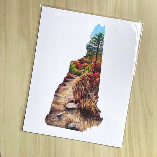 An art print of a nature trail in the shape of New Hampshire. Inside the map outline, a dirt path leads to a pine tree and other brush and forest in fall foliage colors. It is a sunny day with blue sky. Outside the state's shape is page white.
