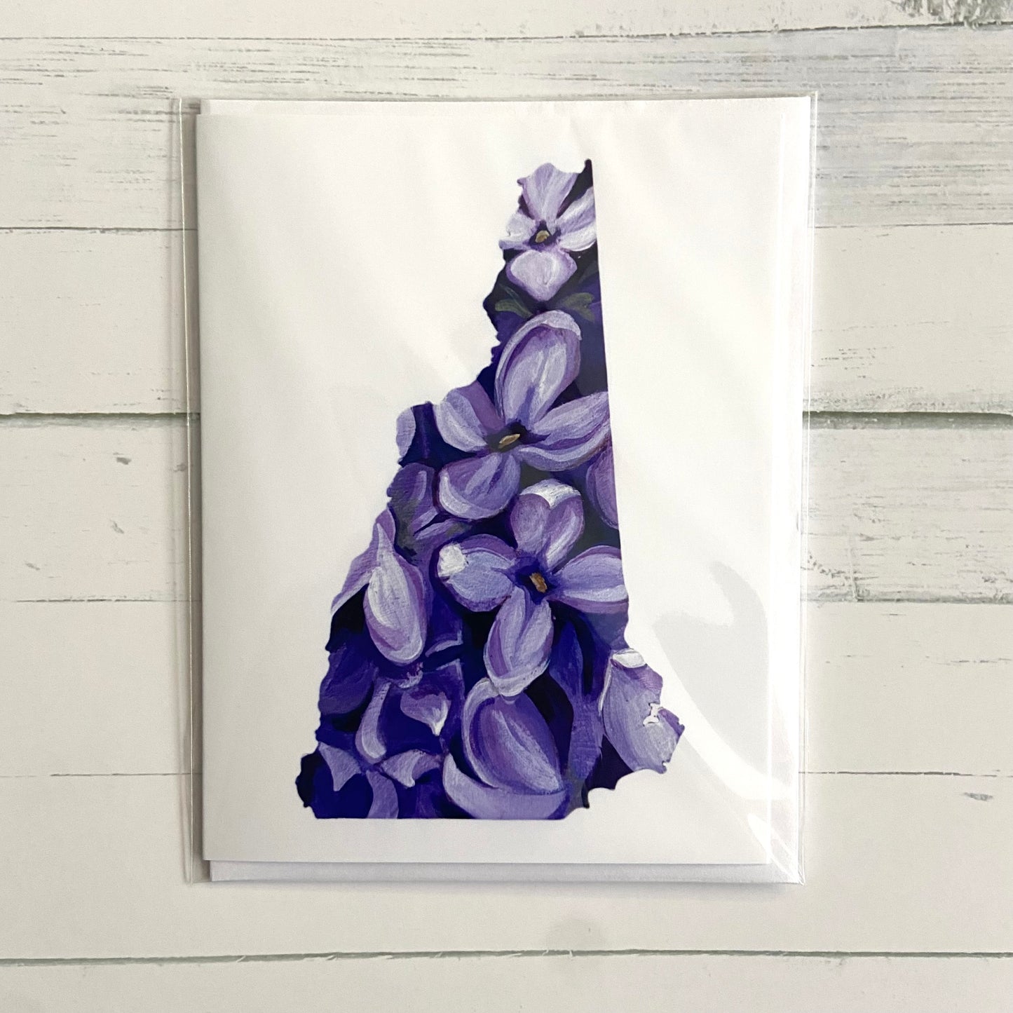 A folded white greeting card with a painting of several purple lilac flowers in the shape of the New Hampshire state outline.