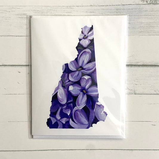 A folded white greeting card with a painting of several purple lilac flowers in the shape of the New Hampshire state outline.