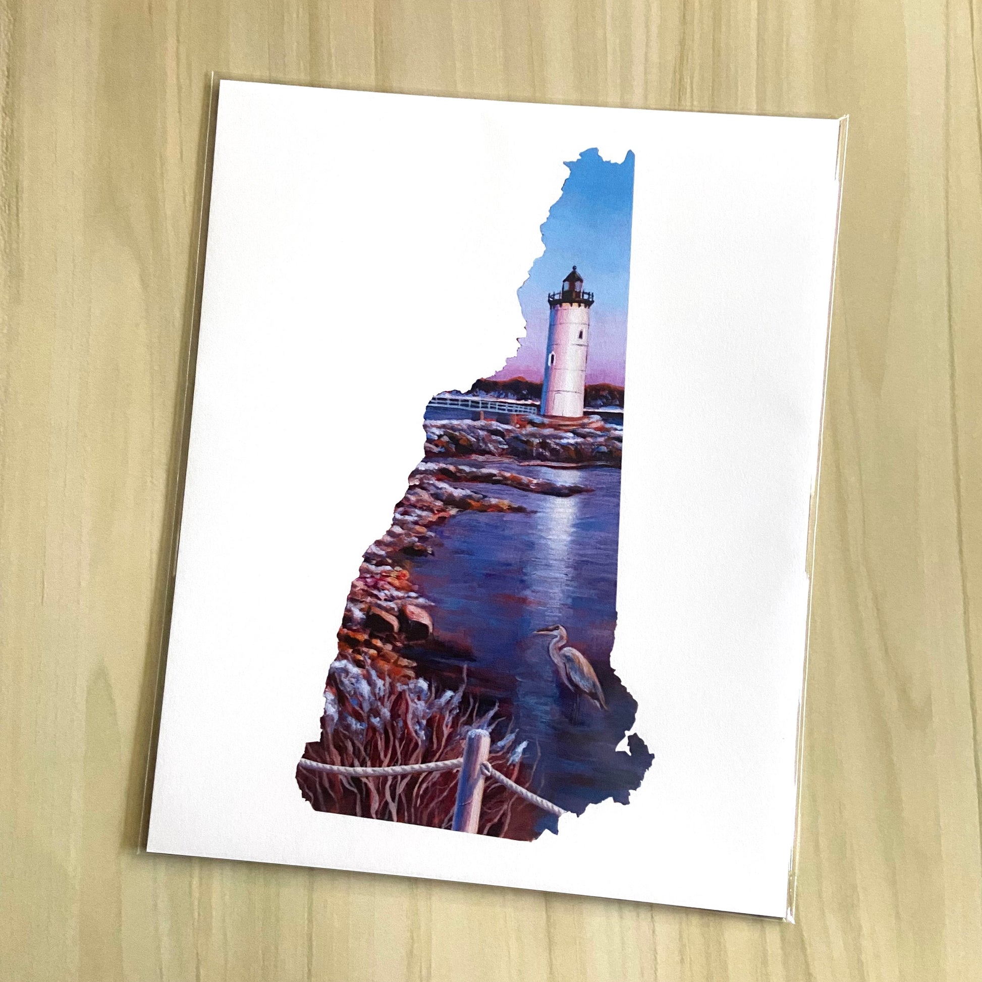 An art print of my painting New Hampshire V sits on a light wood background. The art is in the shape of New Hampshire's map and depicts a lighthouse by a rocky beach with a lone heron with interesting warm lighting.