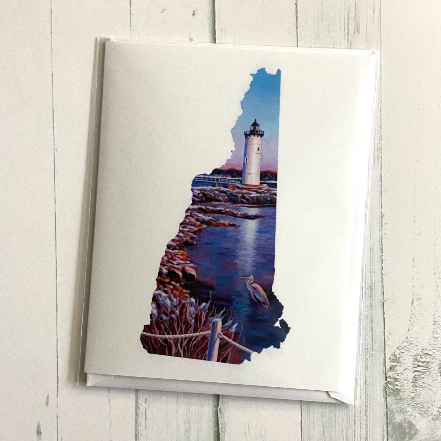 A folded white greeting card with an image of my painting, New Hampshire V, on the front. The art is in the shape of New Hampshire's map and depicts a lighthouse by a rocky beach with a lone heron with interesting warm lighting.