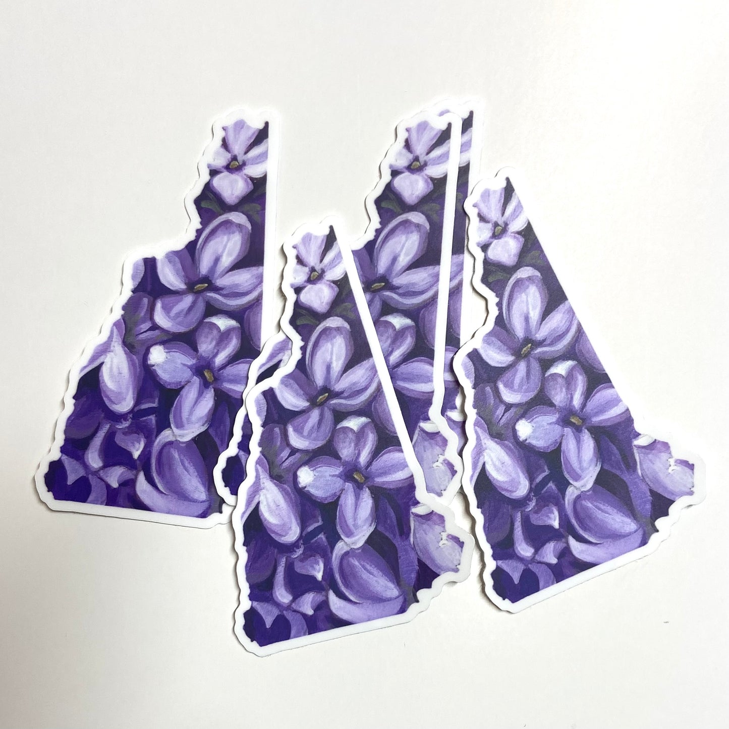 A pile of stickers in the shape of New Hampshire with a painting of purple lilac flowers.