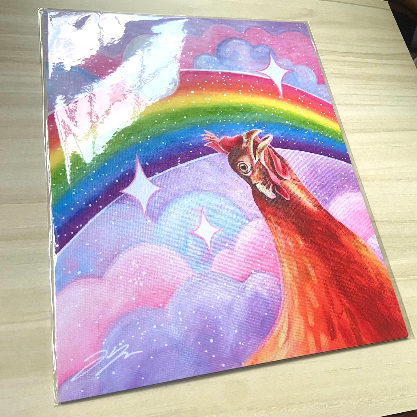 "Nostalgia Hen" print by artist Jackie Hanson, shown packaged as a print on a light wood background. The print shows an orange chicken looking in awe at a vibrant, glittery rainbow in a pink and purple sky.