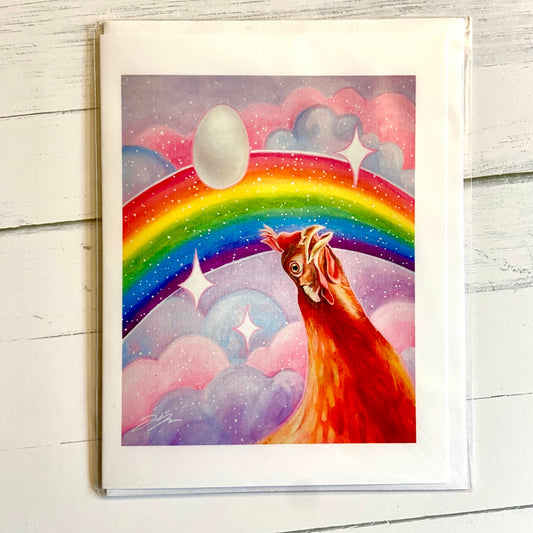 A folded white greeting card with an image of my acrylic painting, Nostalgia Hen. A chicken gazes upward with an expression of wonder toward a pink and purple sky, bisected by a glittering rainbow. A single white egg floats above.