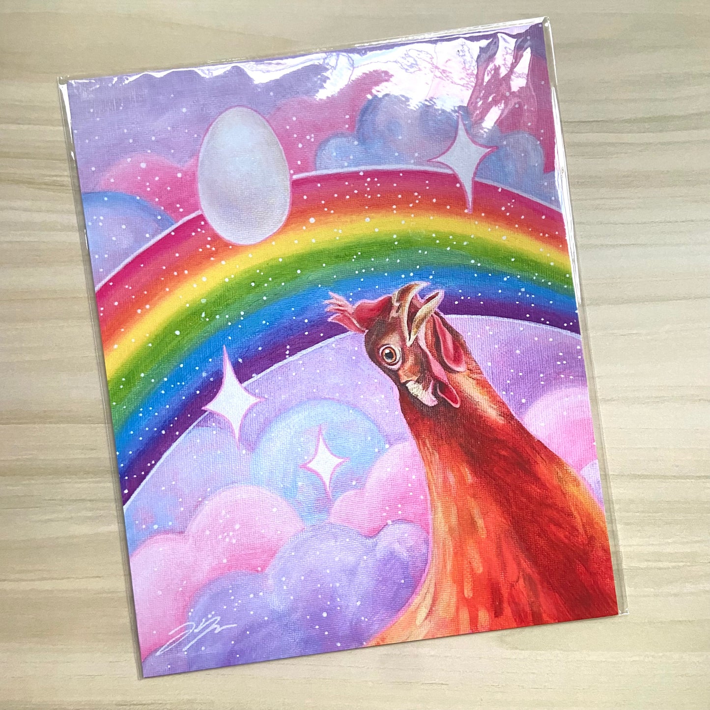 "Nostalgia Hen" print by artist Jackie Hanson, shown packaged as a print on a light wood background. The print shows an orange chicken looking in awe at a vibrant, glittery rainbow in a pink and purple sky.
