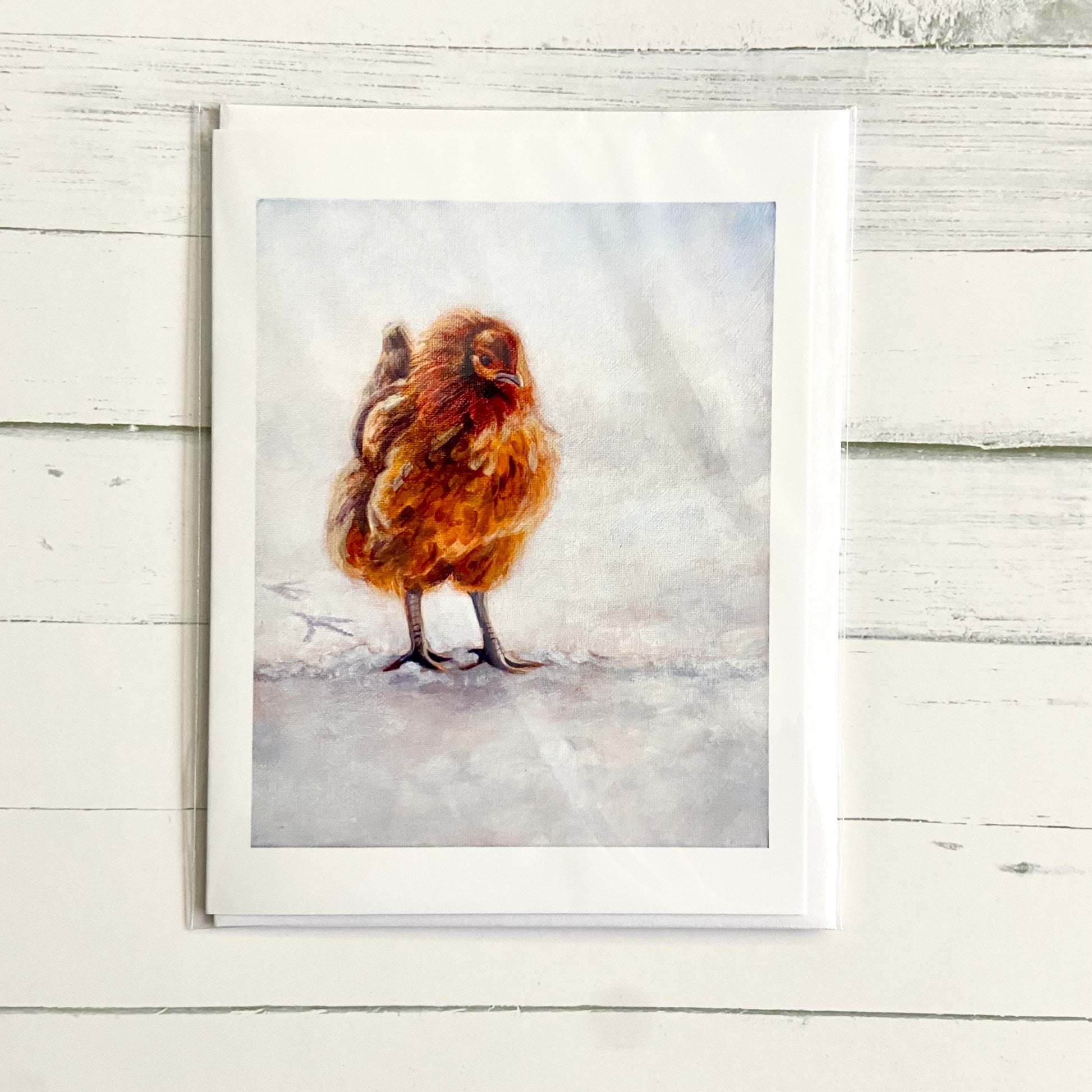 A folded white greeting card with a painting on the front of an orange chicken standing in snow.