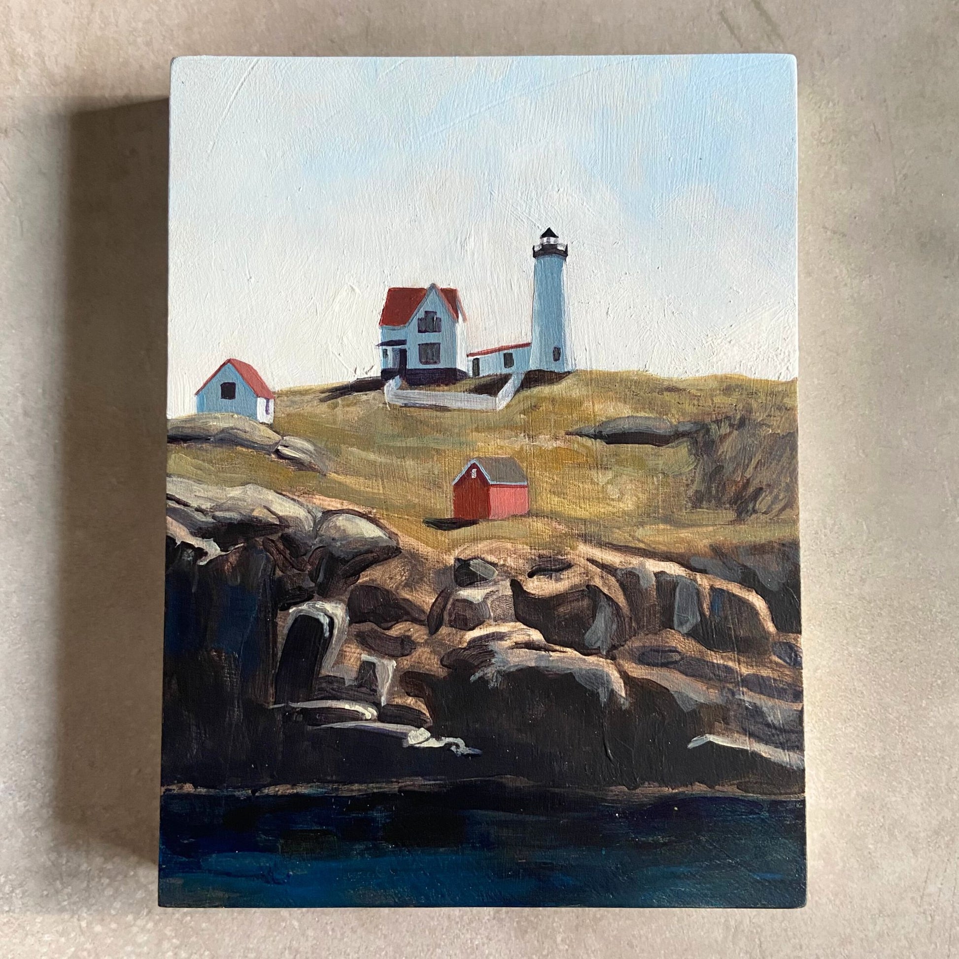 An acrylic painting on wood panel sitting on a beige, textured background. The artwork, portrait-oriented, shows a lighthouse on a green piece of land under a blue sky. The land ends in rocky cliffs with deep blue water below.