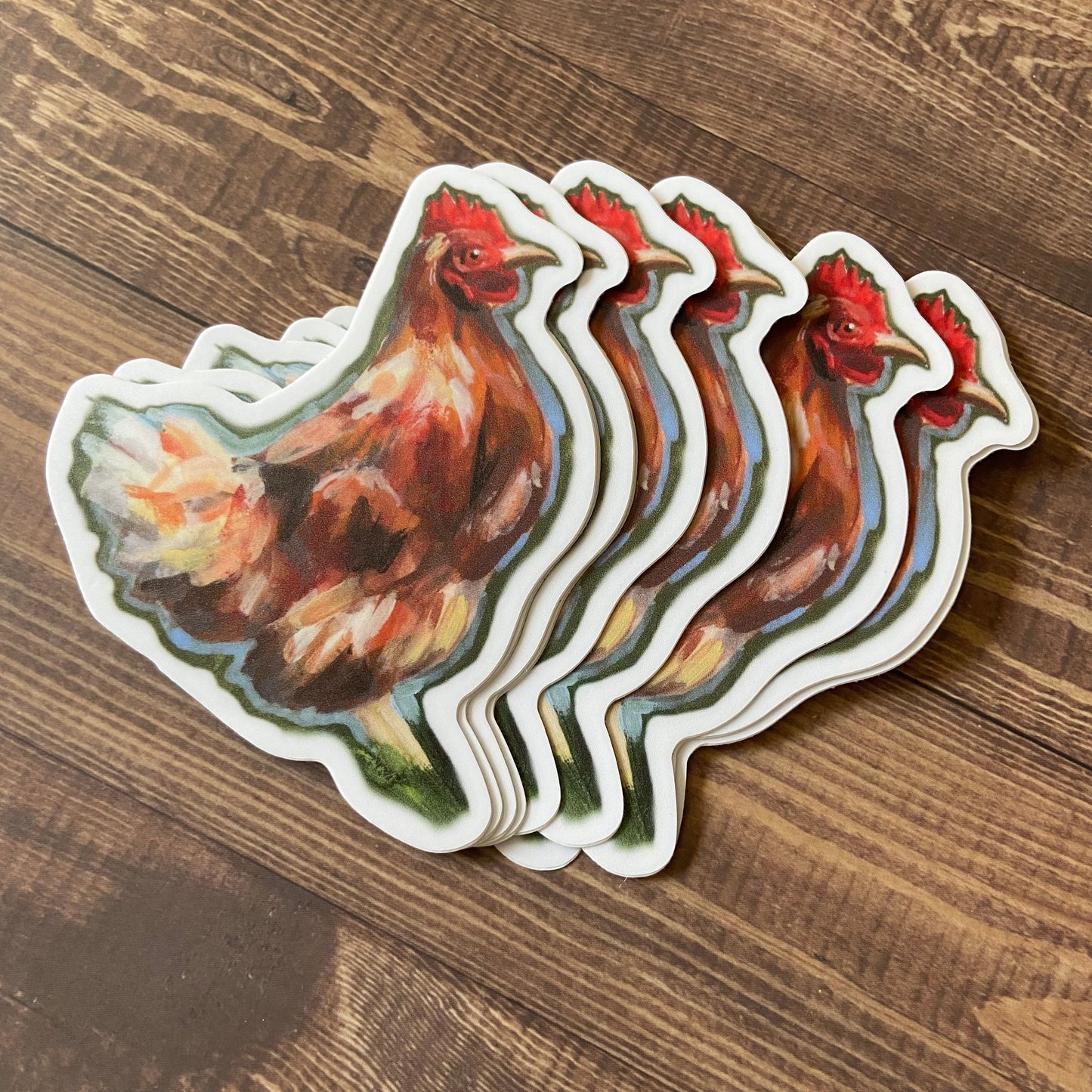 A small pile of orange chicken stickers on a wooden surface. Each sticker is die-cut to hen shape and has a narrow white border. The chickens are clearly painted and have a neutral expression.