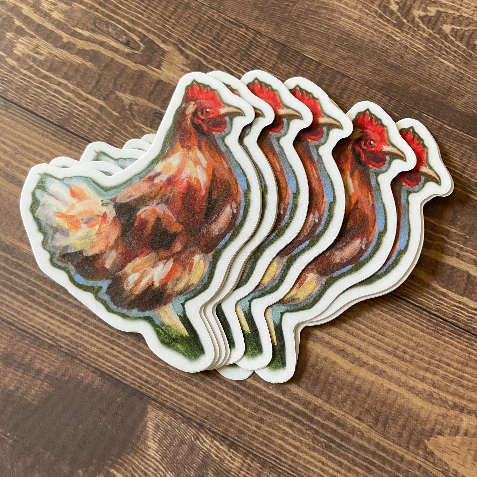 A small pile of orange chicken stickers on a wooden surface. Each sticker is die-cut to hen shape and has a narrow white border. The chickens are clearly painted and have a neutral expression.