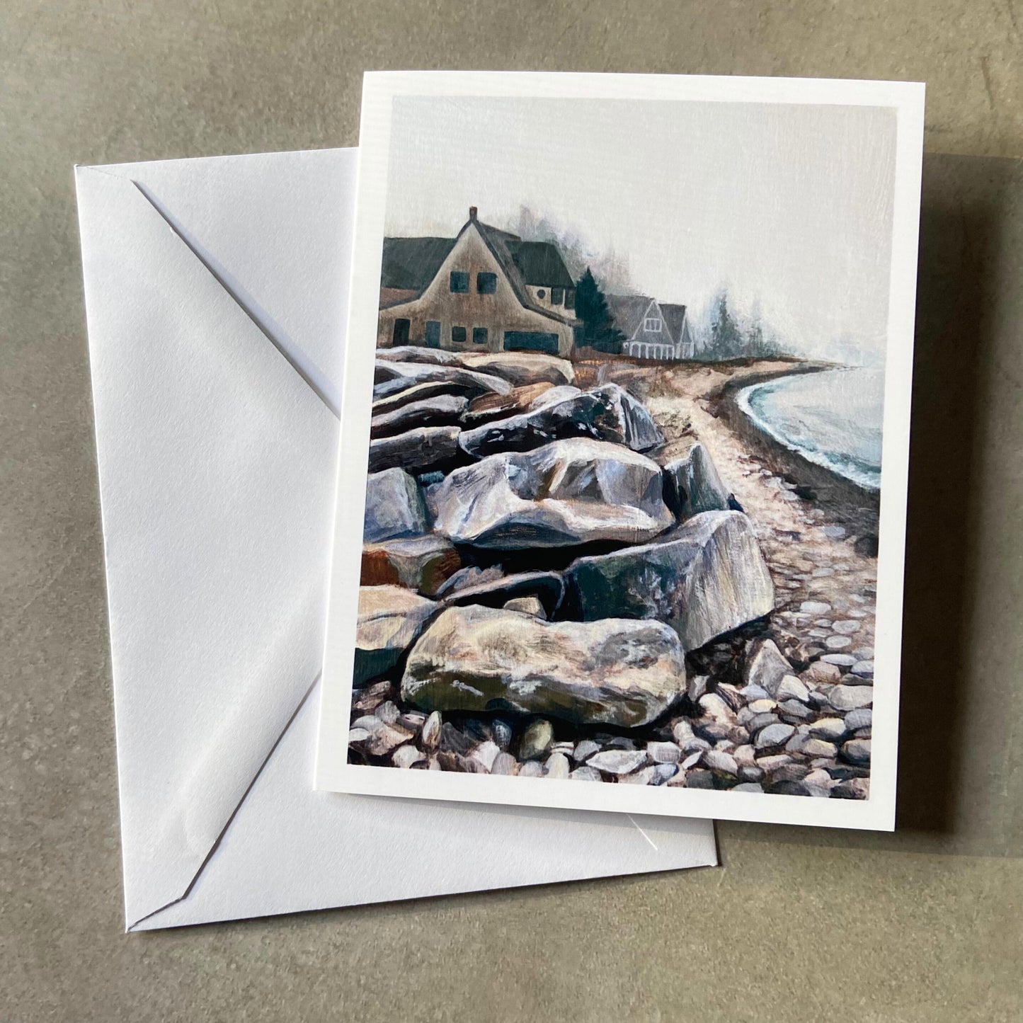 A folded white greeting card and envelope on a grey surface. Depicted on the card is a rocky beach, with a stack of large rocks at left in the foreground. In the background is a row of beach houses disappearing into a foggy day.