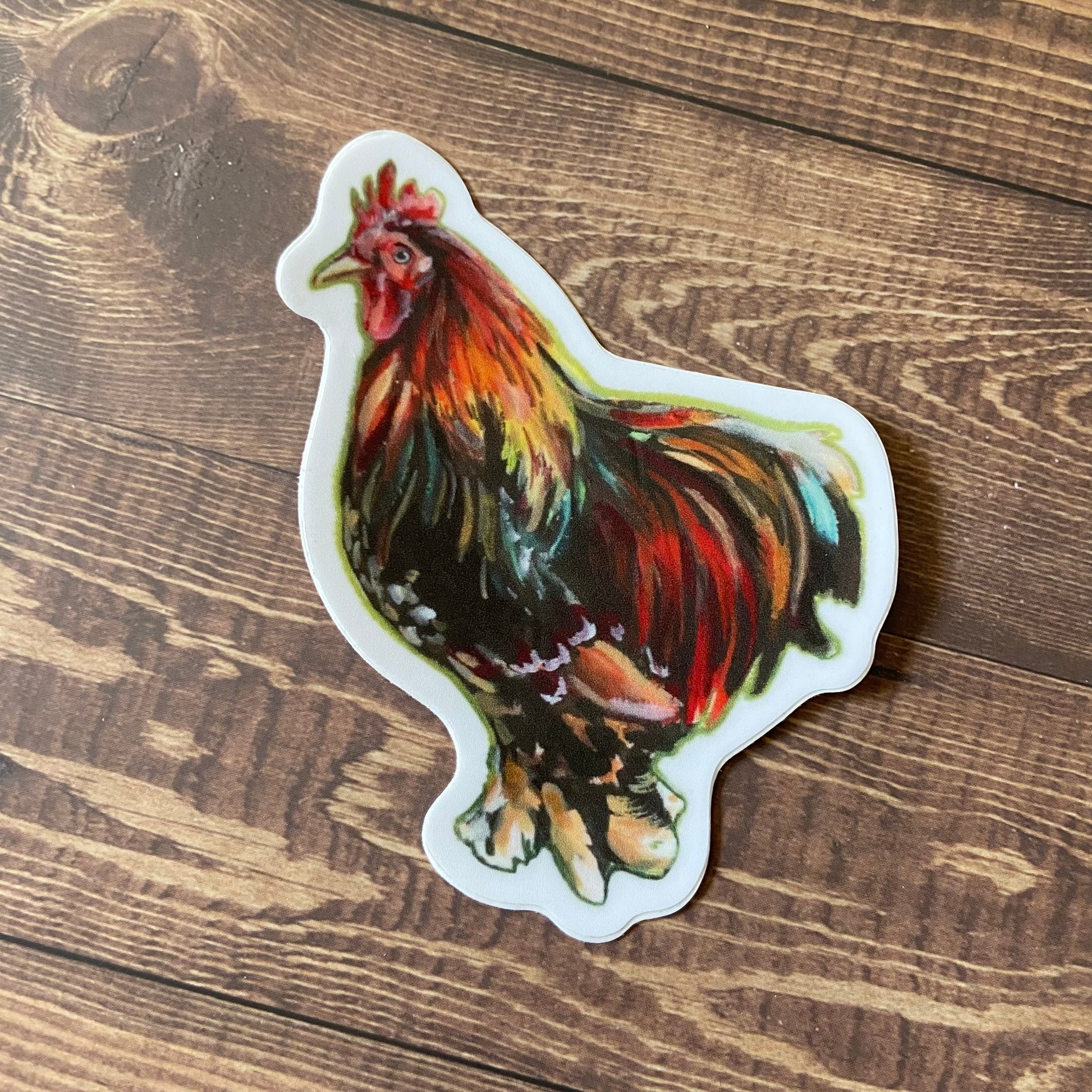 A sticker of a rooster sitting on a wooden surface. The decal shows a painting of a colorful, mature rooster in profile, with delicate brush strokes making each feather. The sticker is die-cut to rooster shape with a thin white border all around.