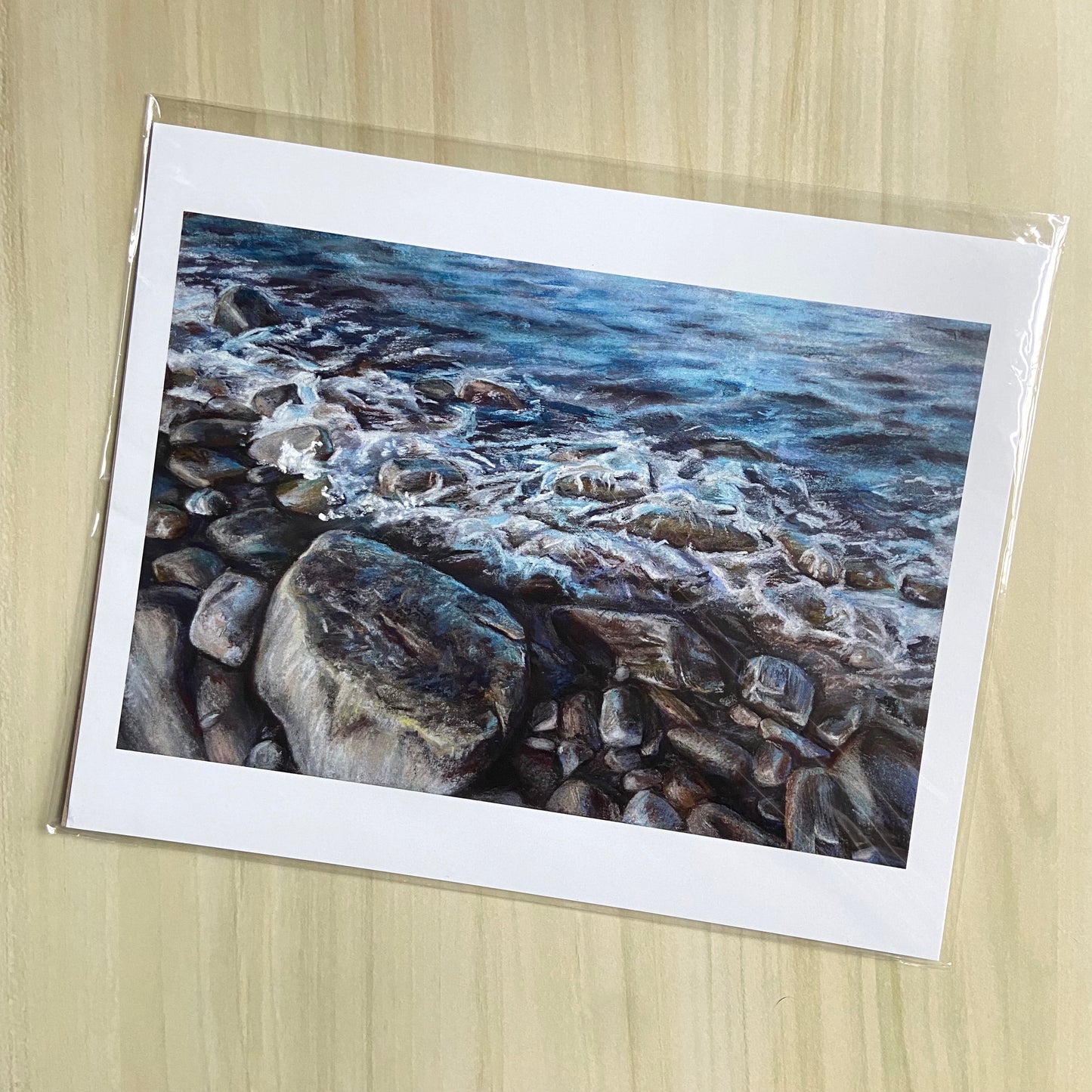 An art print of blue ocean waves crashing onto a rocky beach. The artwork is cropped in close so water fills the top half and stones the bottom. The grainy texture of pastels is shown in this painting's print. There are white borders around the art.
