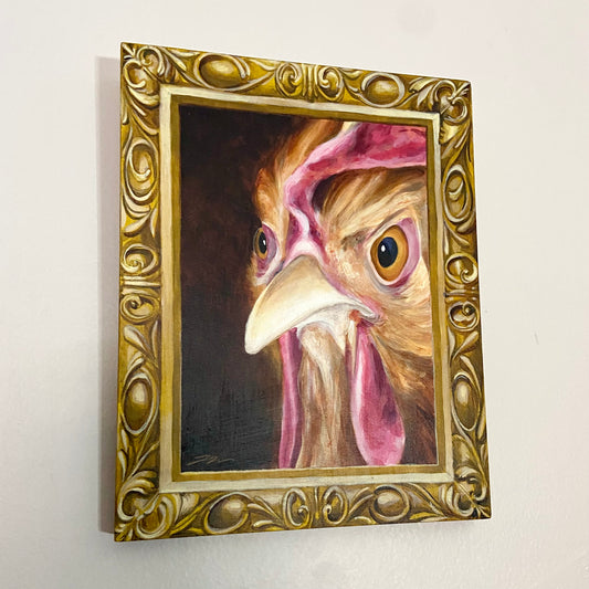 A painting of a chicken's angry face in an ornate gold frame, also painted. The artwork hangs on a light gray wall.