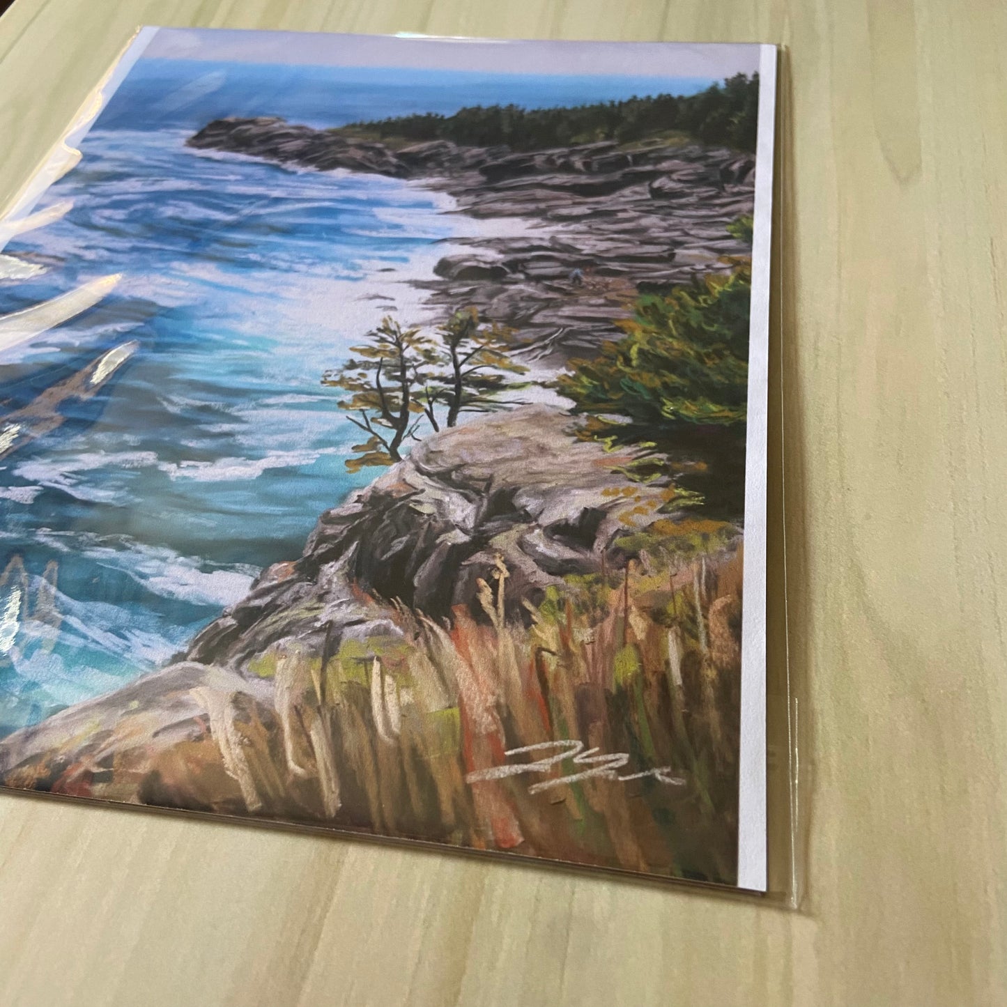 An angled detail view of the Maine coastal art print. The soft pastels used to paint are vibrant and textured, as seen by the dry brush near the bottom. The ocean is bright blue with swirling waves and the rocks on the beach are rugged and gray.