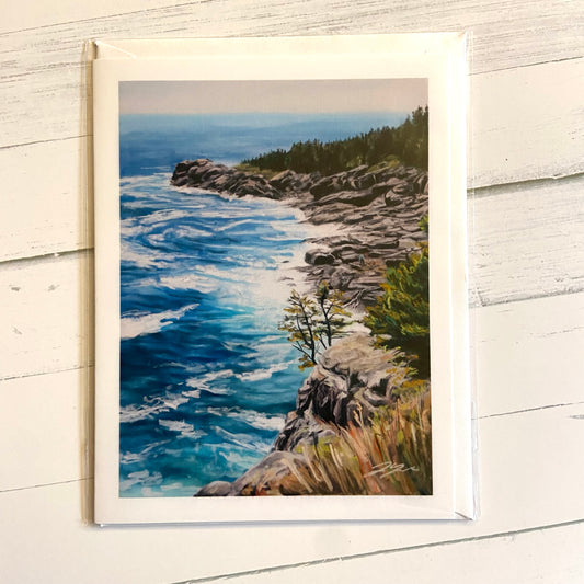 A folded white greeting card with a painting of a rocky Maine cliff and beach meeting the bright blue sea.