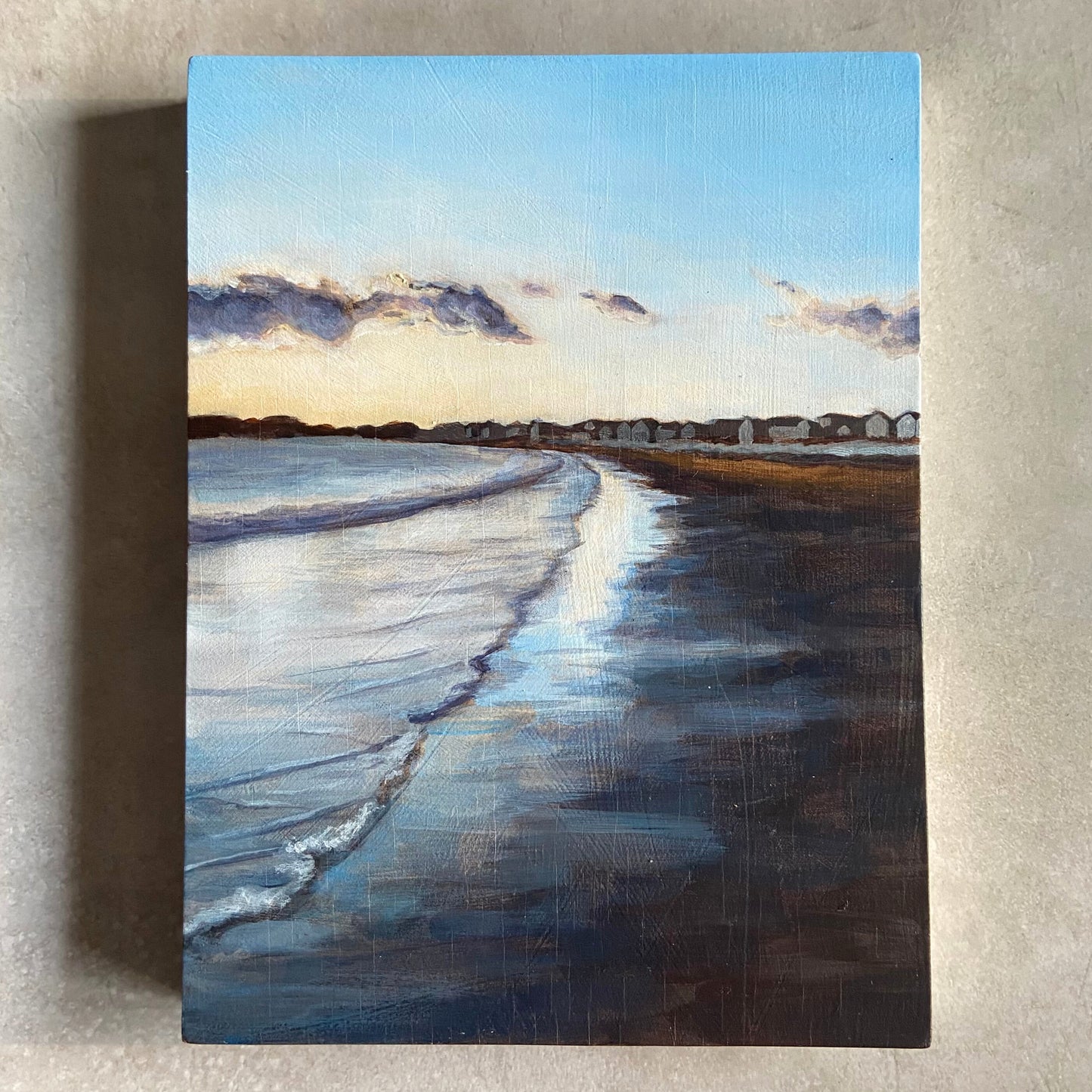 An acrylic seascape painting on wood panel, sitting on a beige, textured background. The painting shows a sand beach at sunset, with gentle waves meeting the wet sand and both reflecting the evening sky. On the horizon is a row of beachfront homes.