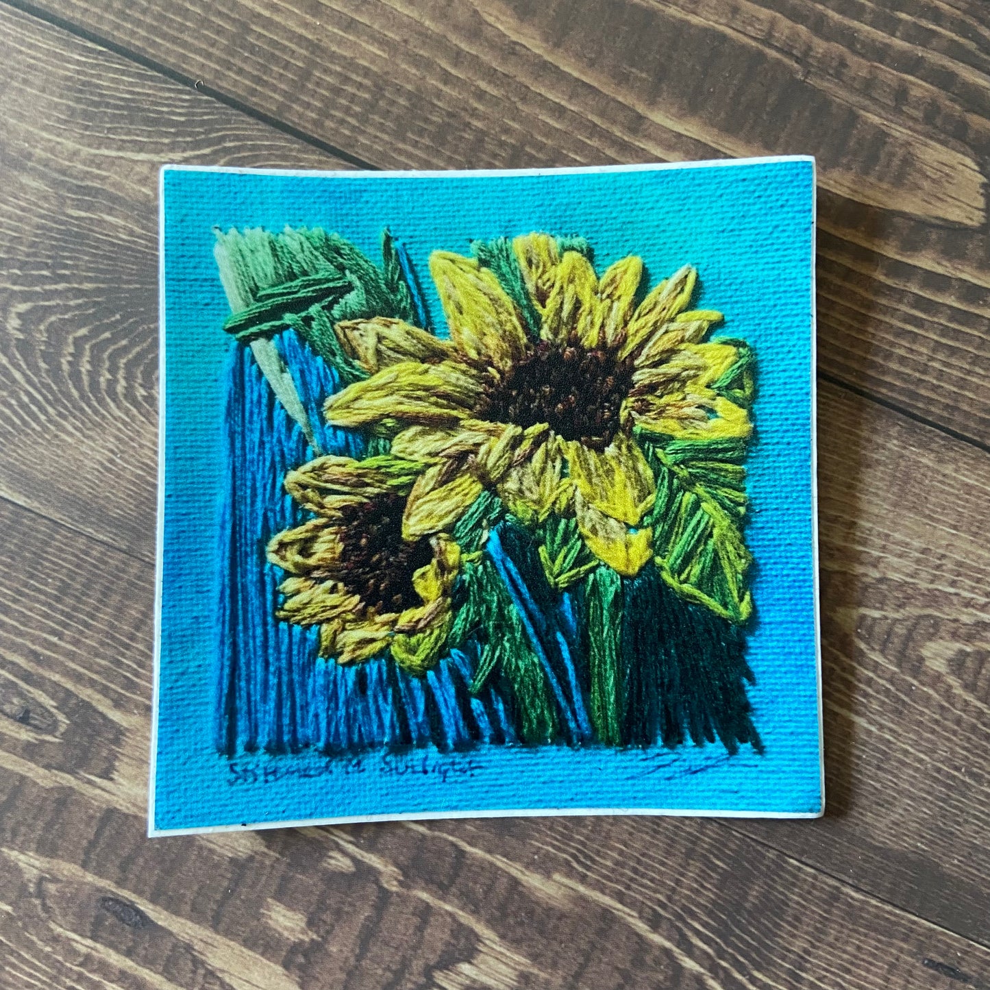 A square sticker of an embroidered painting of two sunflowers. The texture of the canvas and the texture of the thread are very clear. The painting is primarily light blue with the sunflowers in vibrant yellow.