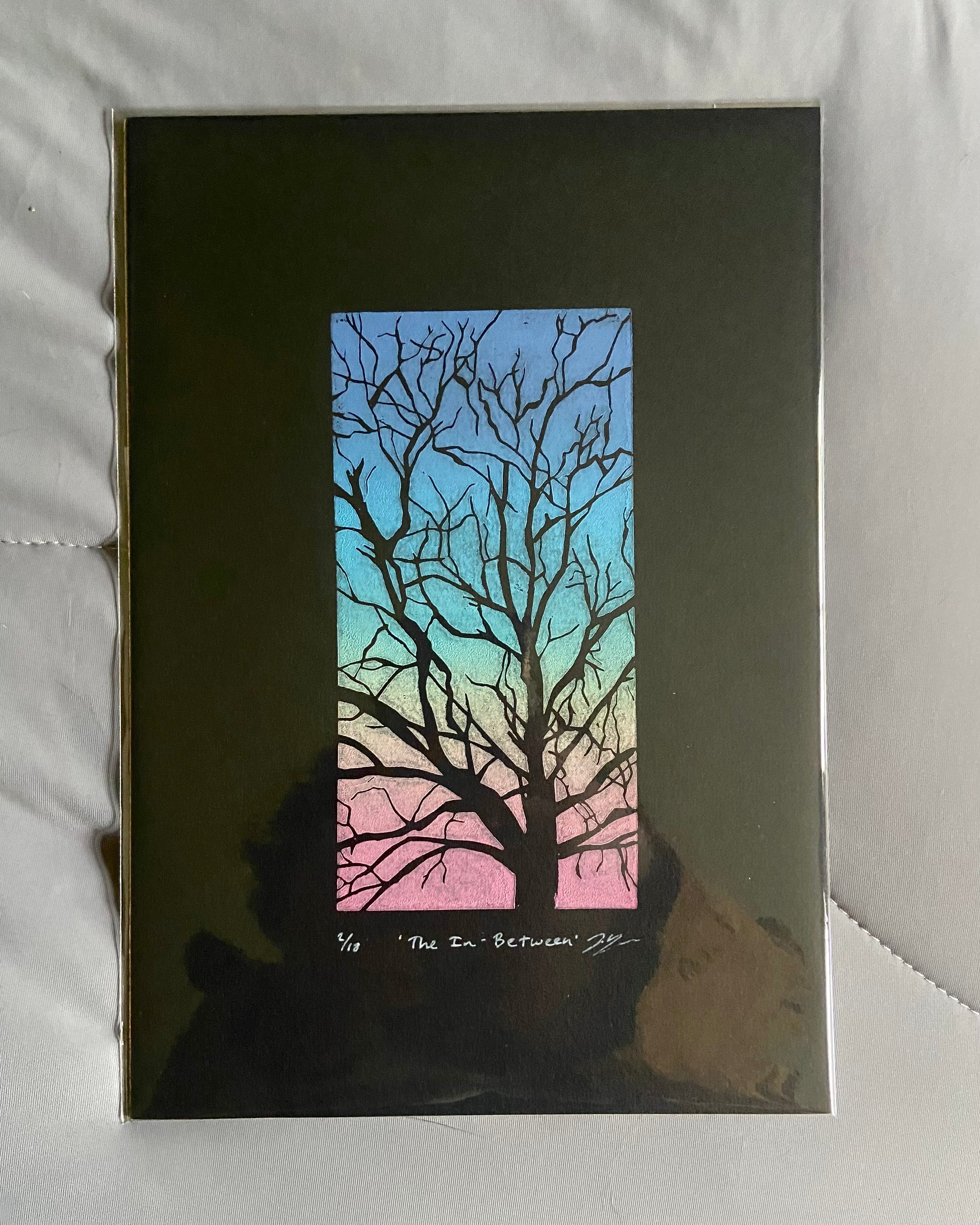 The relief print as packaged. Black tree branches are shown between negative space printed in a rainbow gradient in a 3x6 vertical space. Written in white below is the edition number, title "The In-Between", and the artist's signature.