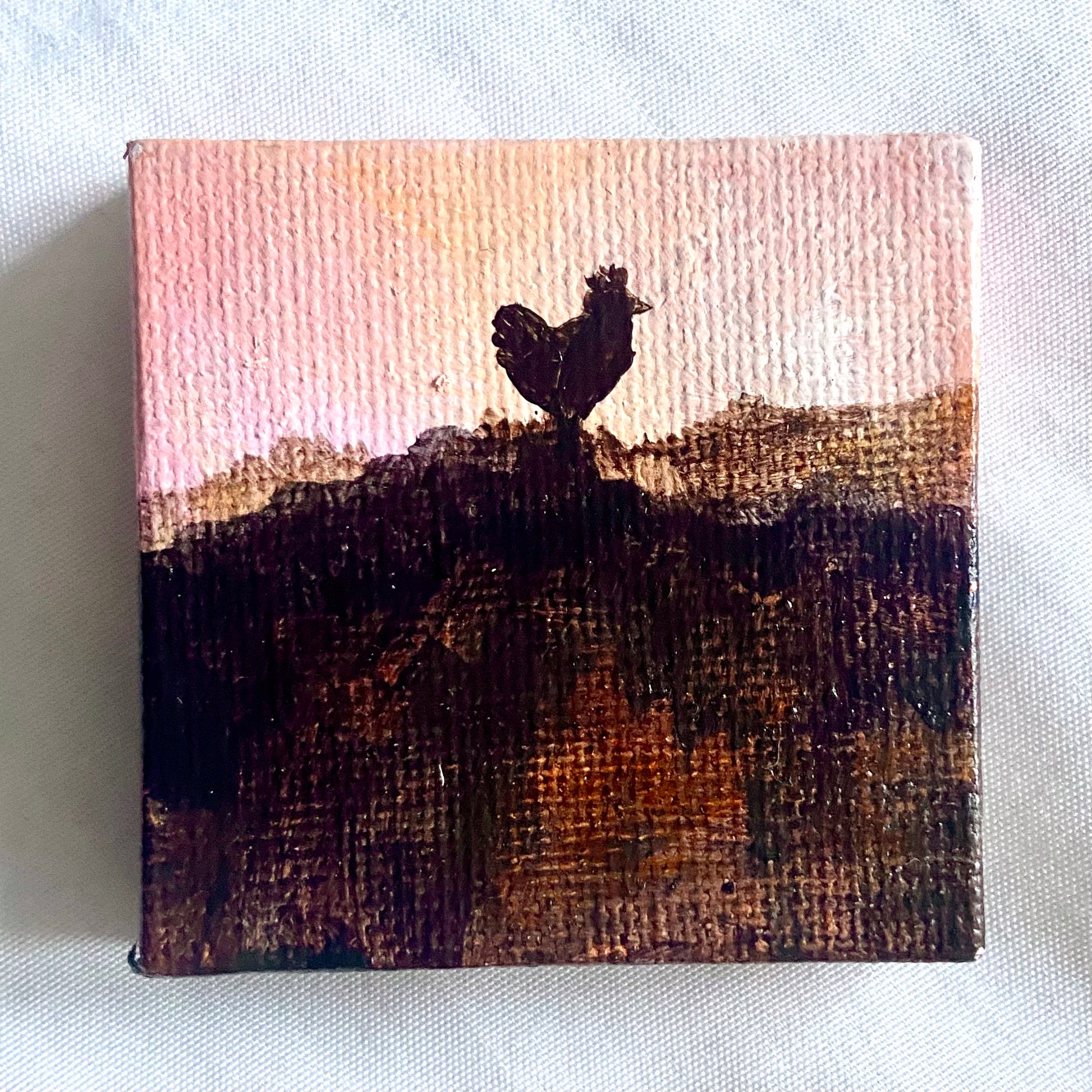 A small square painting of a chicken in silhouette, on top of a dark hill in front of a light pink sky.