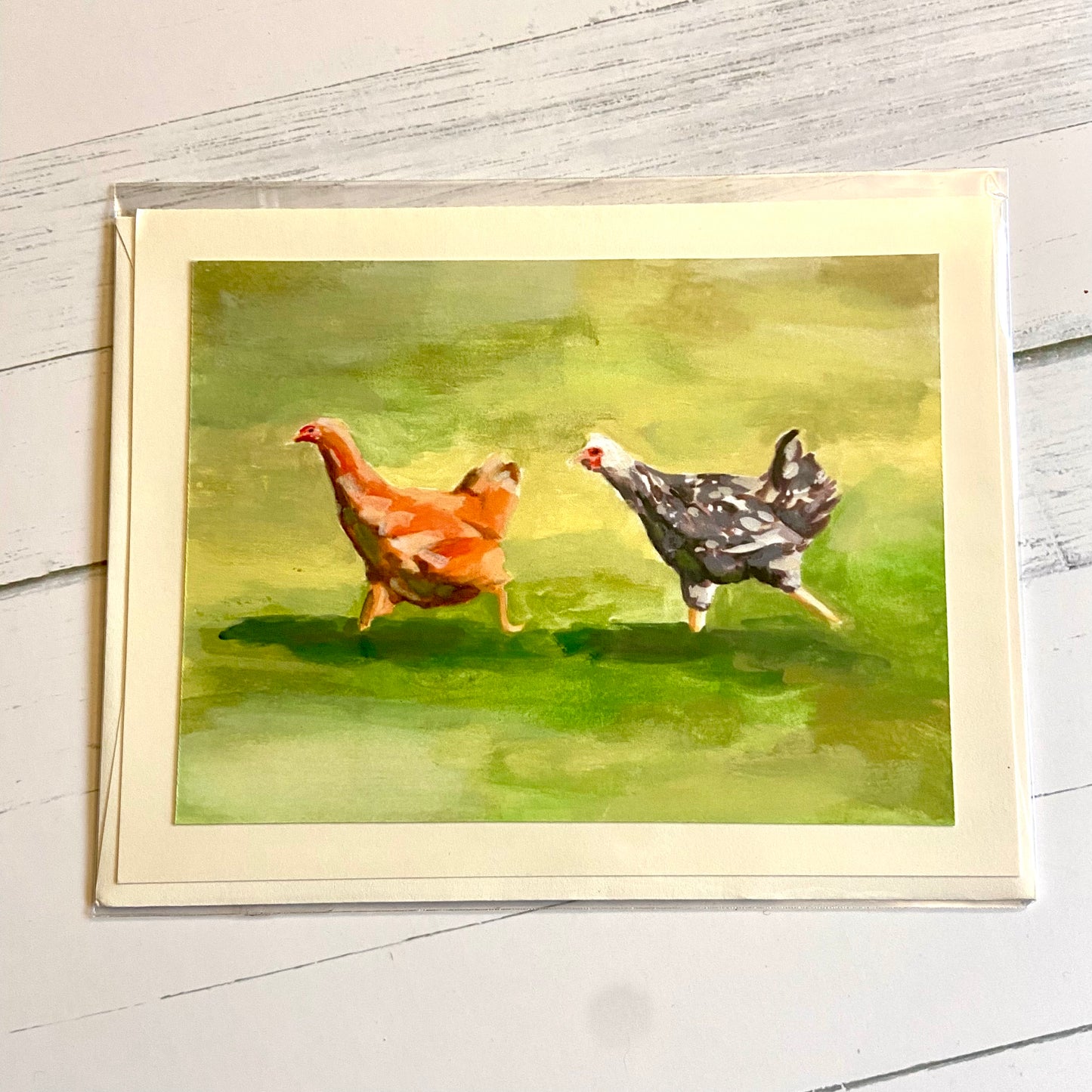 A folded white greeting card depicting ym gouache painting, Two Running Hens. Depicted are an orange and a black-and-white hen, both mid-stride in a sprint across a green lawn.