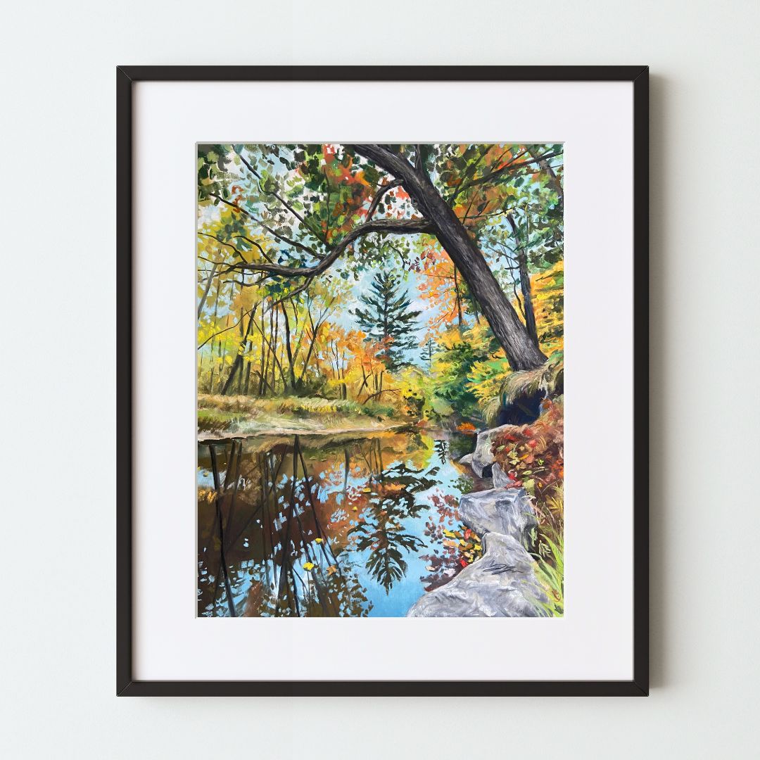 A mockup of a landscape art print in a black frame with a white mat. A still, reflective river runs through a forest during peak fall foliage. A tree near the viewer on the right leans out over the river, its branches overhead.